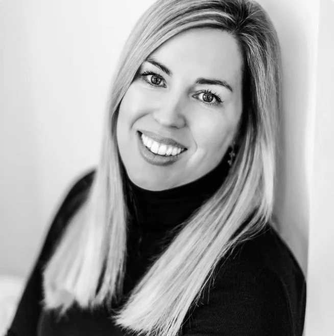 Black and white portrait of a smiling woman with straight blonde hair, wearing a dark turtleneck and earrings.