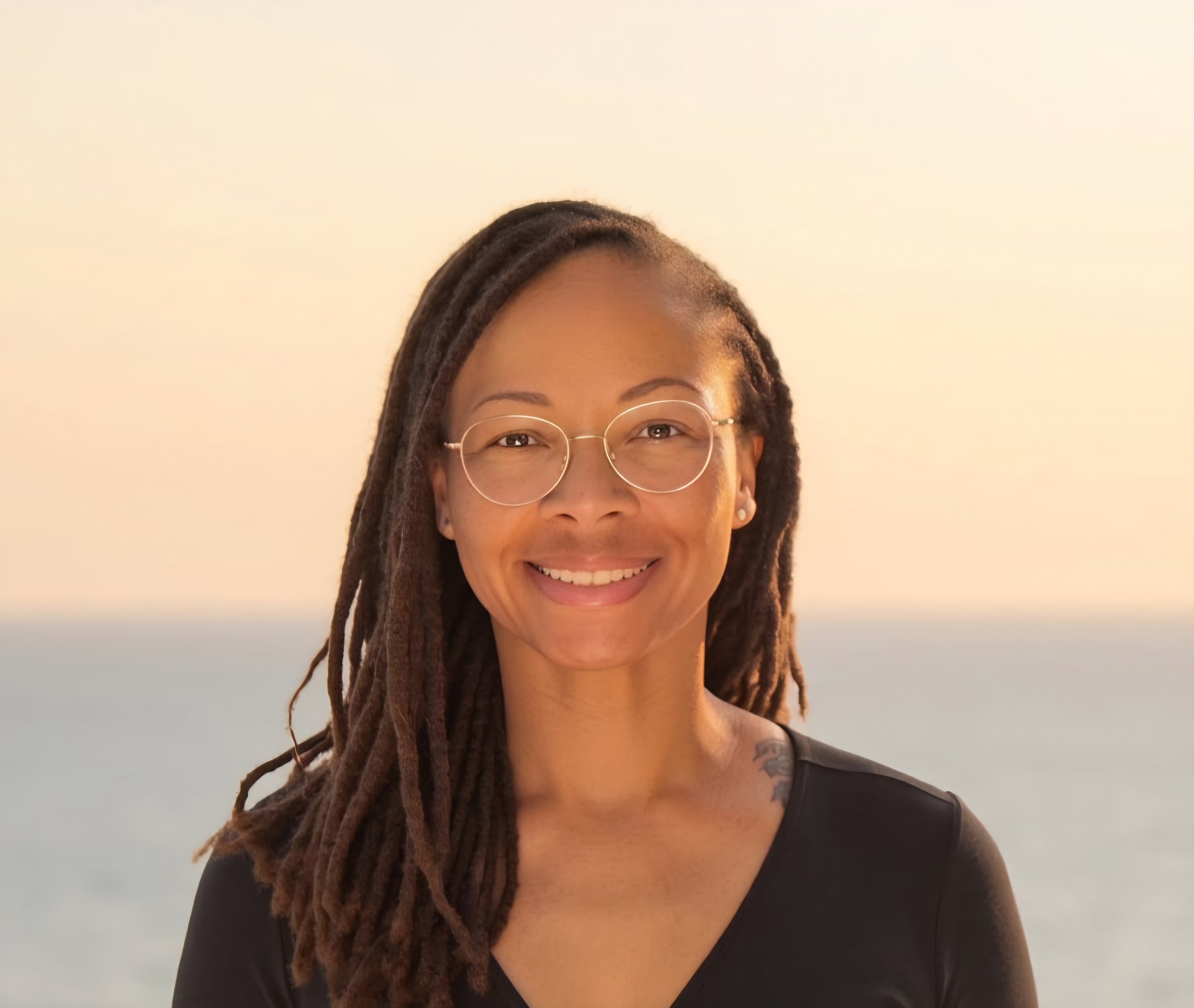 A woman smiling with long dreadlocks, wearing glasses and a black top, with a light-colored background.