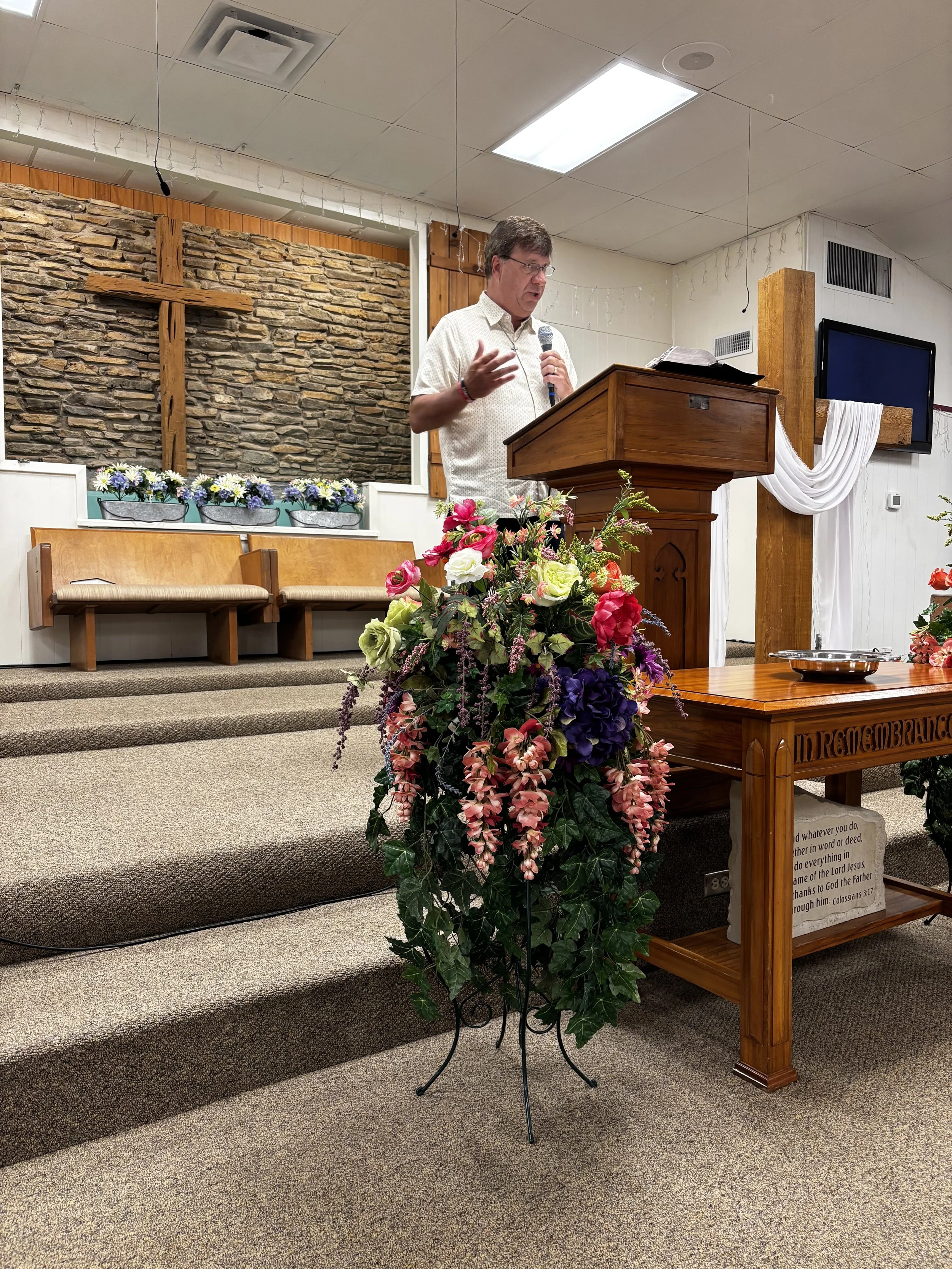 A man speaking at a church pulpit decorated with a large floral arrangement, with an open Bible on the stand, in a room with wood-paneled walls and a stone cross on the wall behind him.