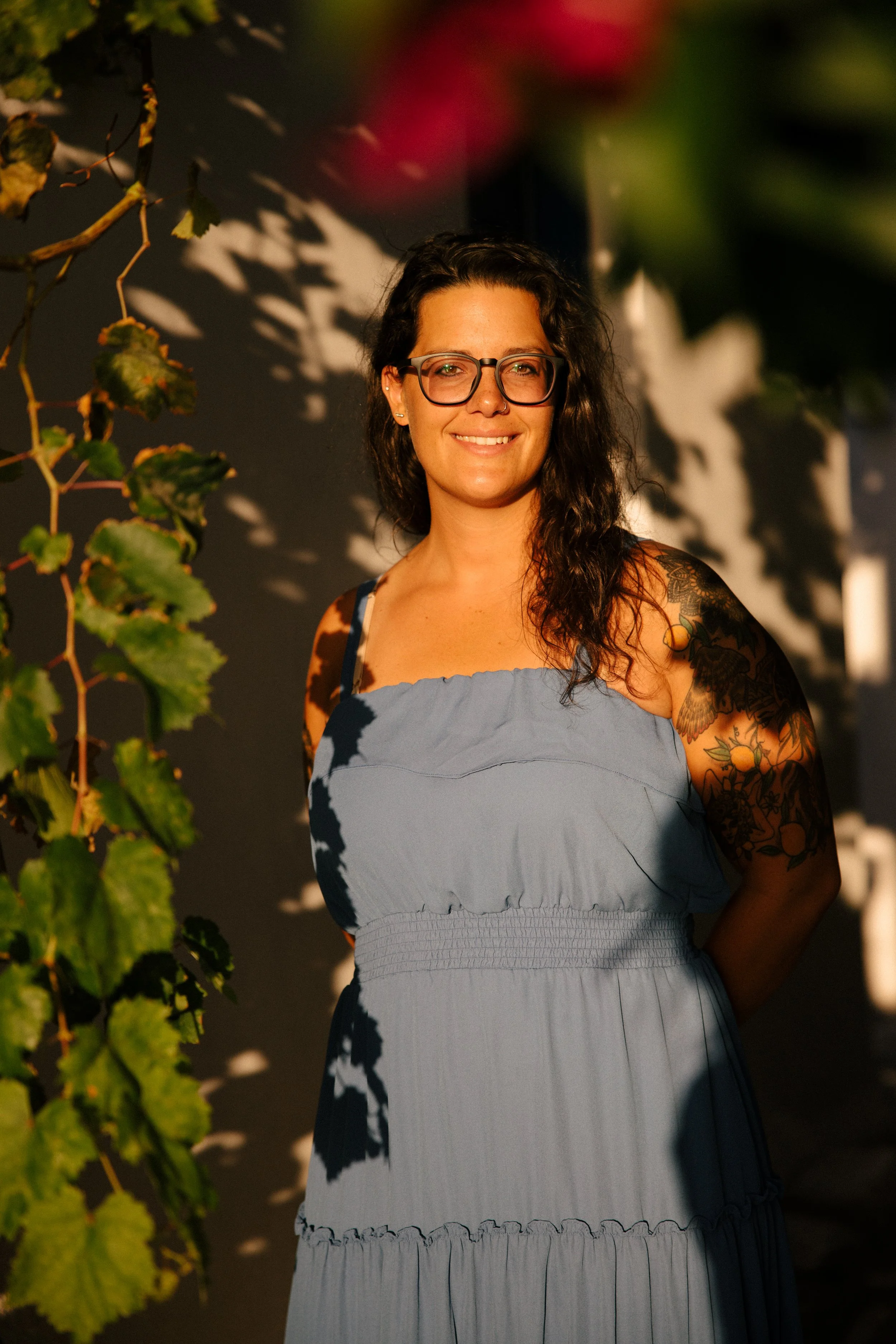 A woman with long dark curly hair, glasses, and tattoos on her arm, wearing a light blue dress, smiling outdoors with leaves and shadows on a dark background.