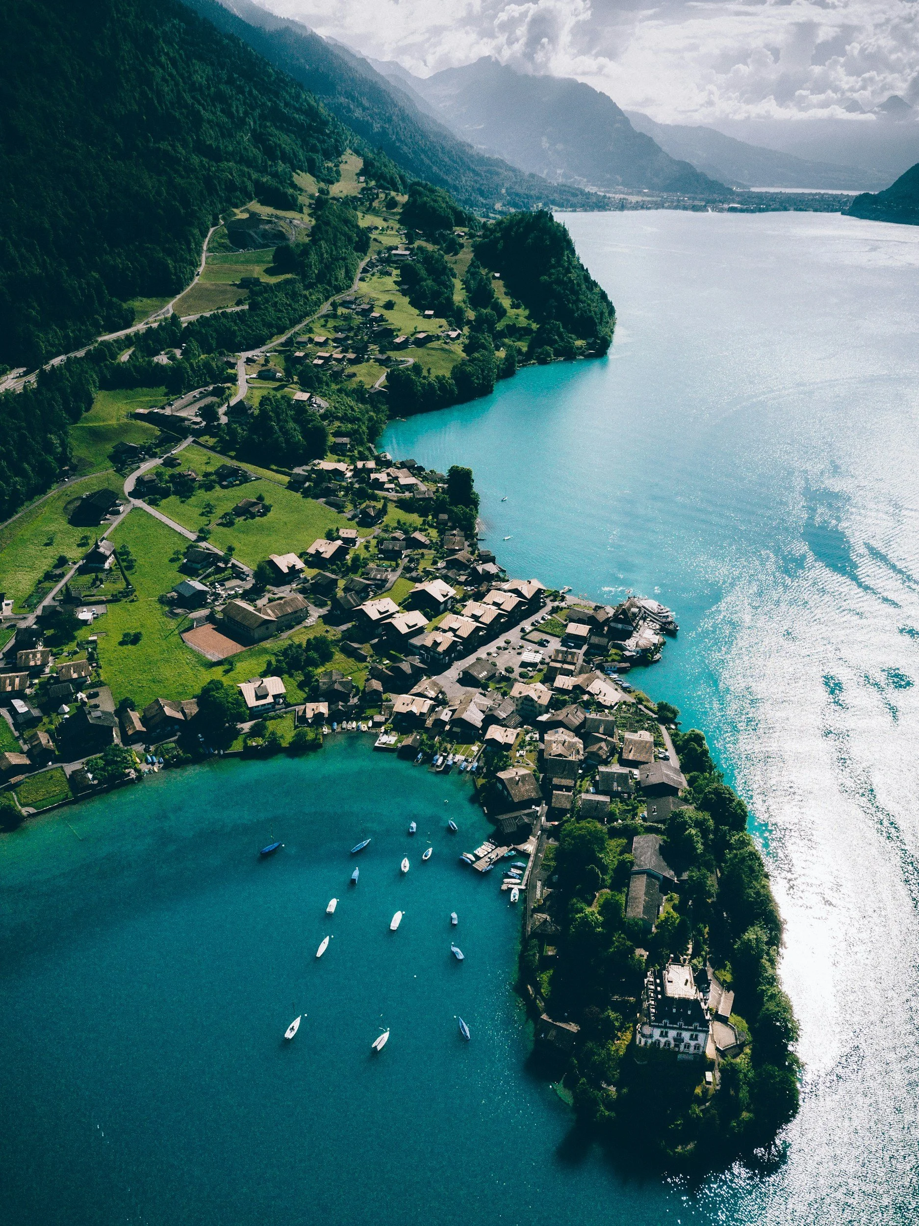 Aerial view of a lakeside village with houses and boats, surrounded by green mountains and water.