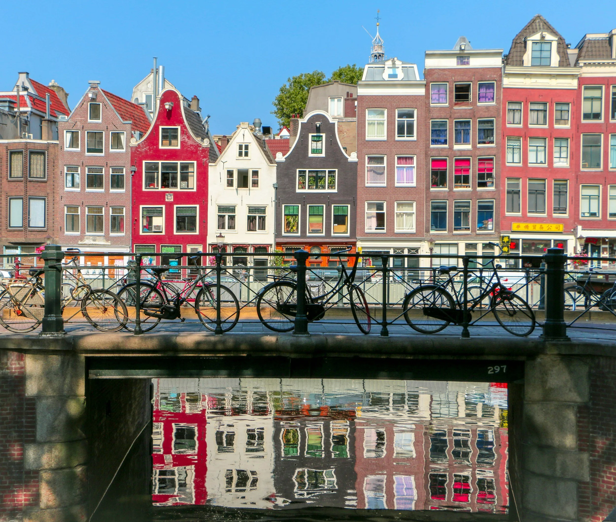 Colorful traditional European buildings with gabled roofs and decorative facades reflect on a canal under a clear blue sky, with bicycles parked along the canal railings.