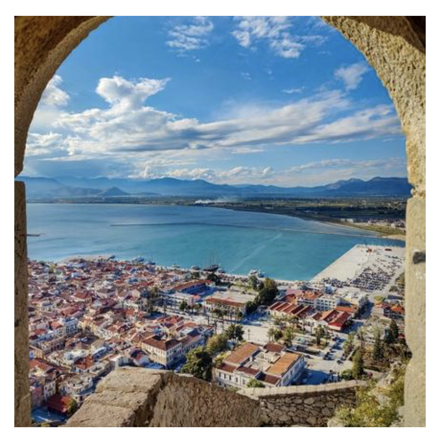 A view of a coastal city through a stone archway, featuring a beach, buildings with red rooftops, and a large body of water with mountains in the background under a partly cloudy sky.