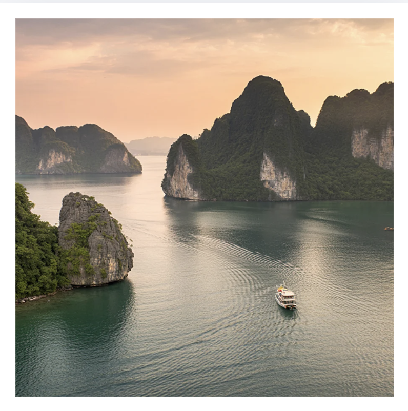 A boat sailing through a calm, greenish waterway surrounded by towering limestone islands covered in lush vegetation, with a sunset sky in the background.