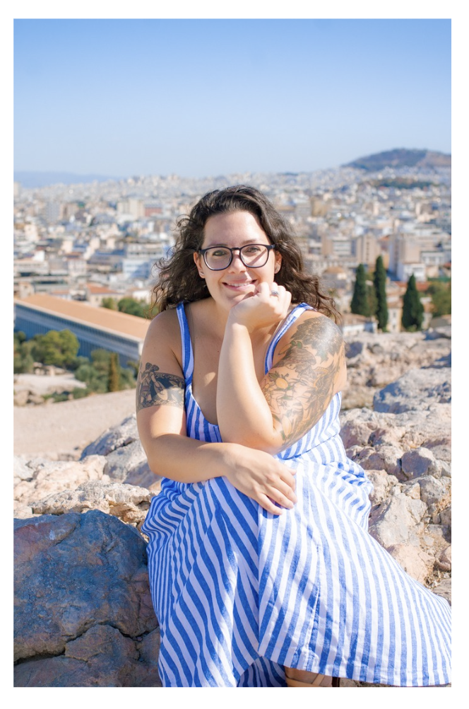 A woman with dark curly hair, glasses, and tattoos on her arms, sitting on rocks outdoors, smiling, with a cityscape and mountainous background under a clear blue sky.