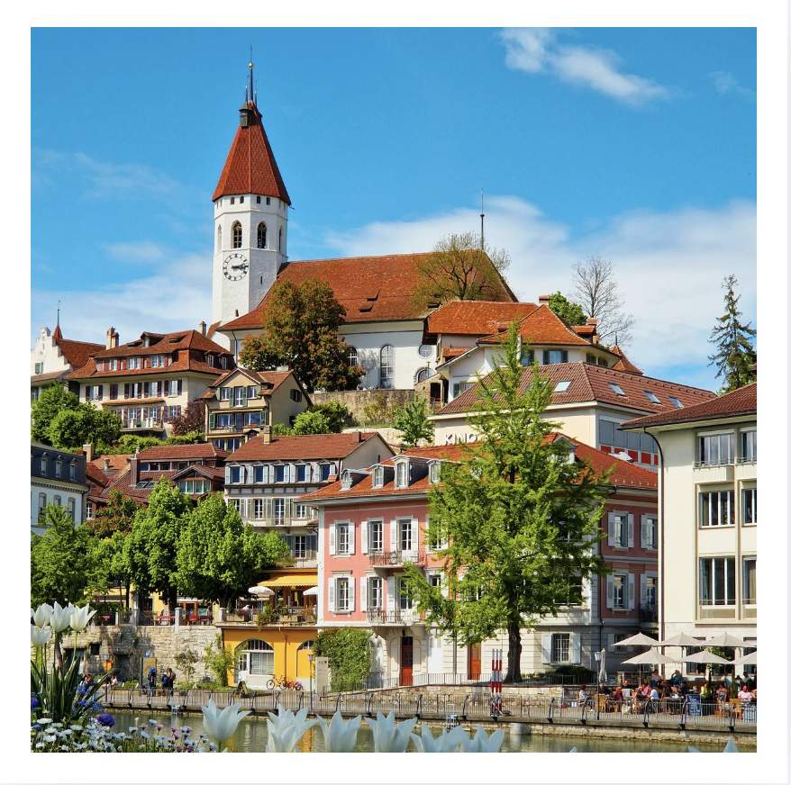 Scenic view of a hillside town with colorful buildings, a church with a tall steeple, trees, a river, and outdoor seating under umbrellas during daytime.