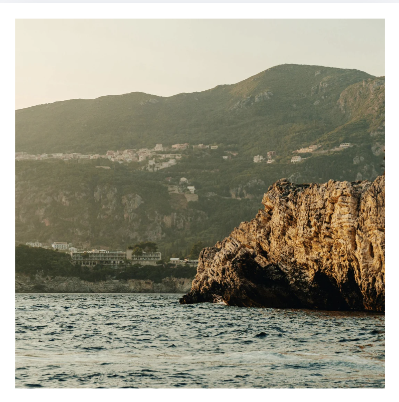 Ocean water with large rocky formation in foreground and green mountainous landscape with houses in the background