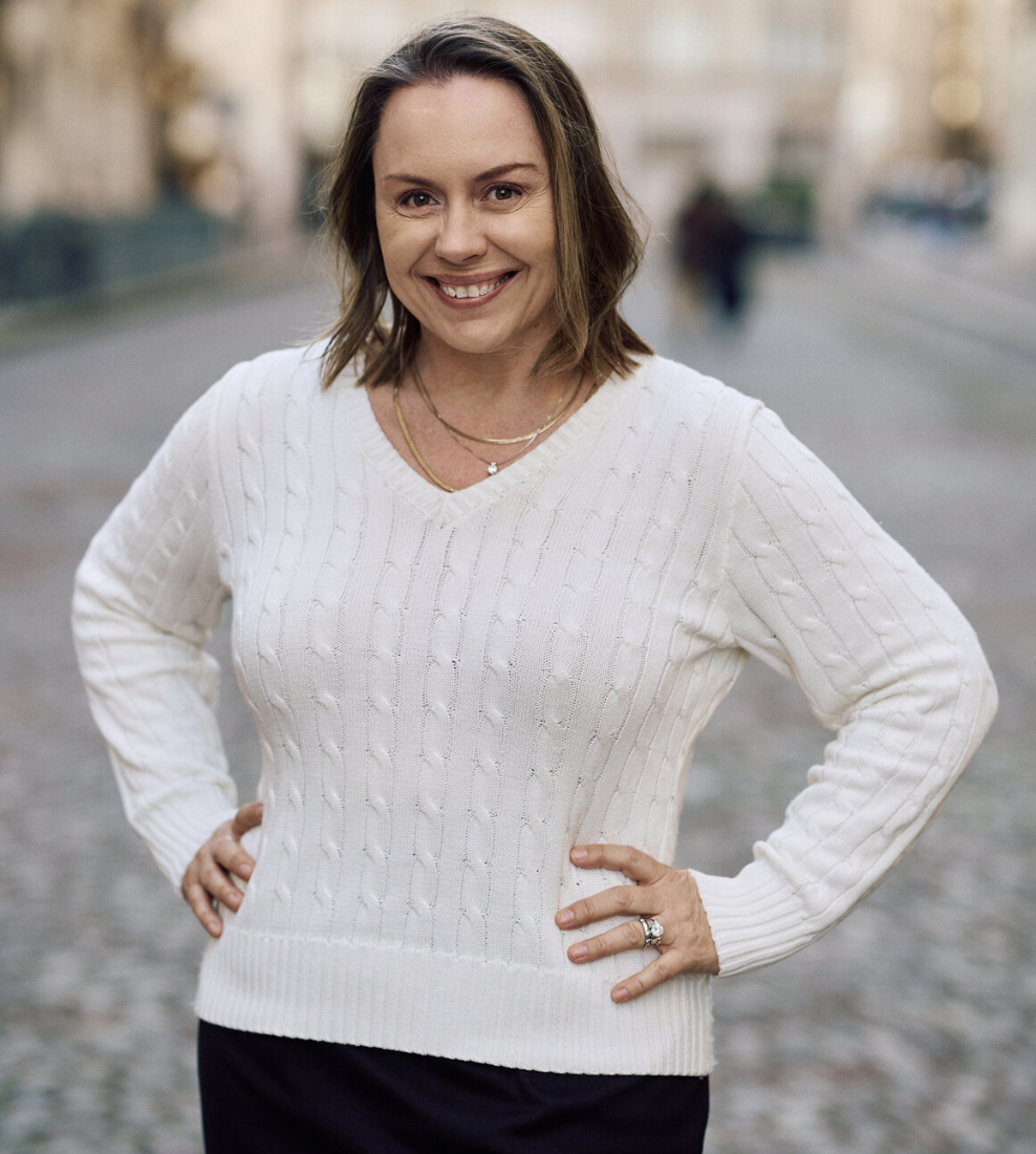 A woman with shoulder-length brown hair, wearing a white sweater and necklaces, smiling with hands on her hips, standing outdoors on a city street.