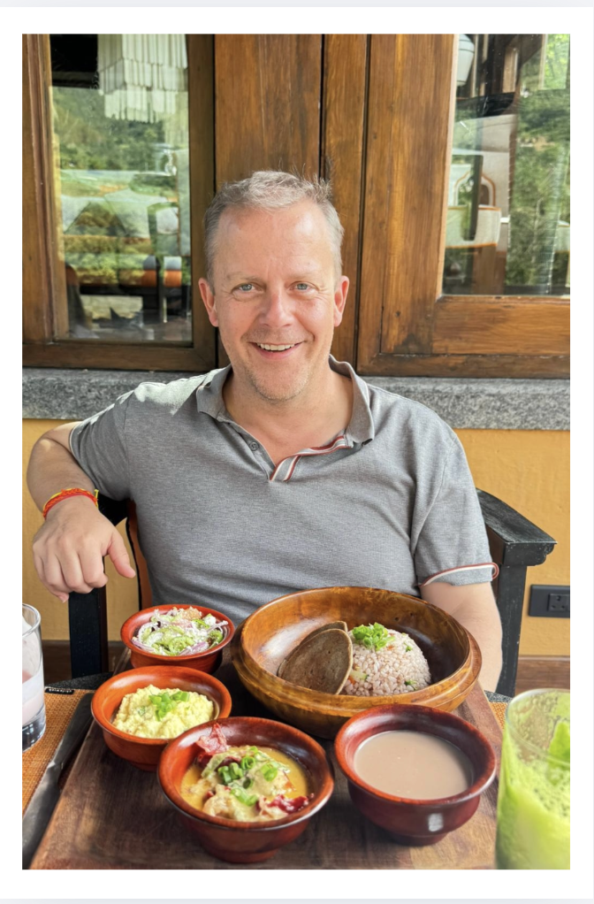 A smiling man with short gray hair wearing a gray polo shirt, sitting at a table with various Mexican dishes in bowls, in a restaurant with wooden walls and windows showing greenery outside.