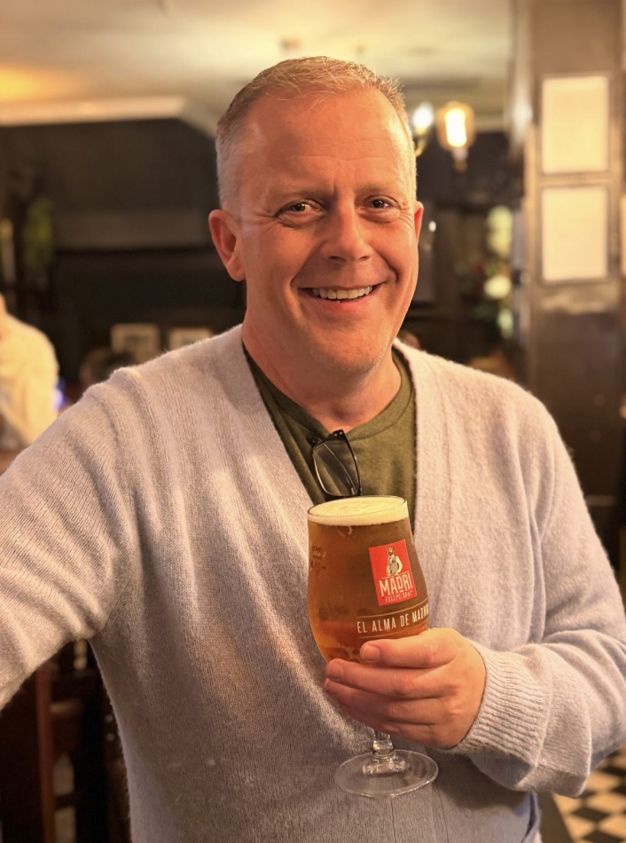 A smiling man holding a glass of beer inside a dimly lit bar or restaurant.