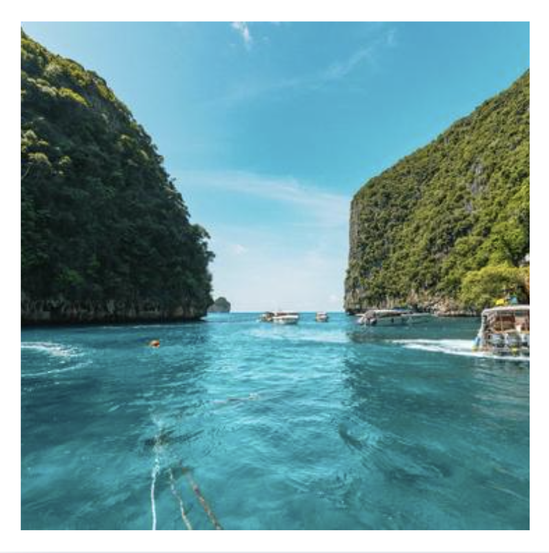 A scenic view of a tropical bay with turquoise water, surrounded by steep, green cliffs on both sides, and boats floating on the water under a clear blue sky.