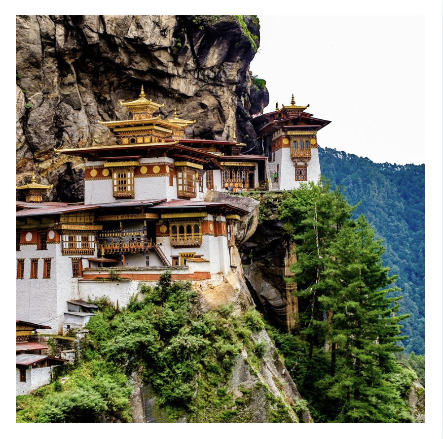 A monastery built into a steep mountainside with traditional Bhutanese architectural style, featuring ornate roofs, white walls, and colorful wooden windows, surrounded by green trees and cliffs.