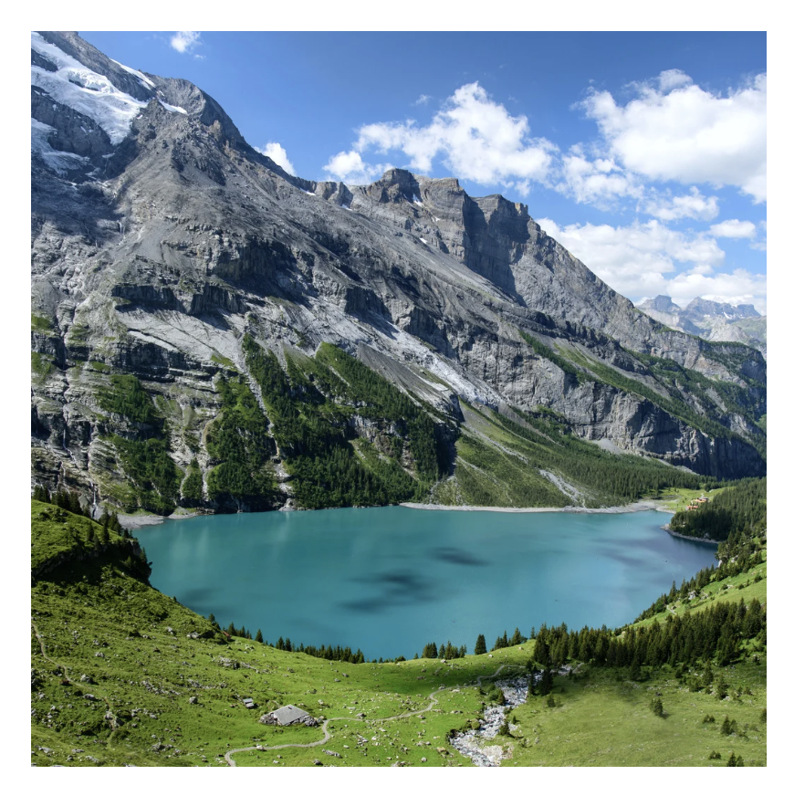 Scenic mountain landscape with a turquoise lake, green meadows, and rocky cliffs under a partly cloudy sky.