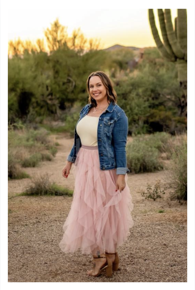 A woman standing outdoors in a desert landscape during sunset, wearing a denim jacket, a light yellow top, a pink tulle skirt, and tan high-heeled sandals, and smiling at the camera.