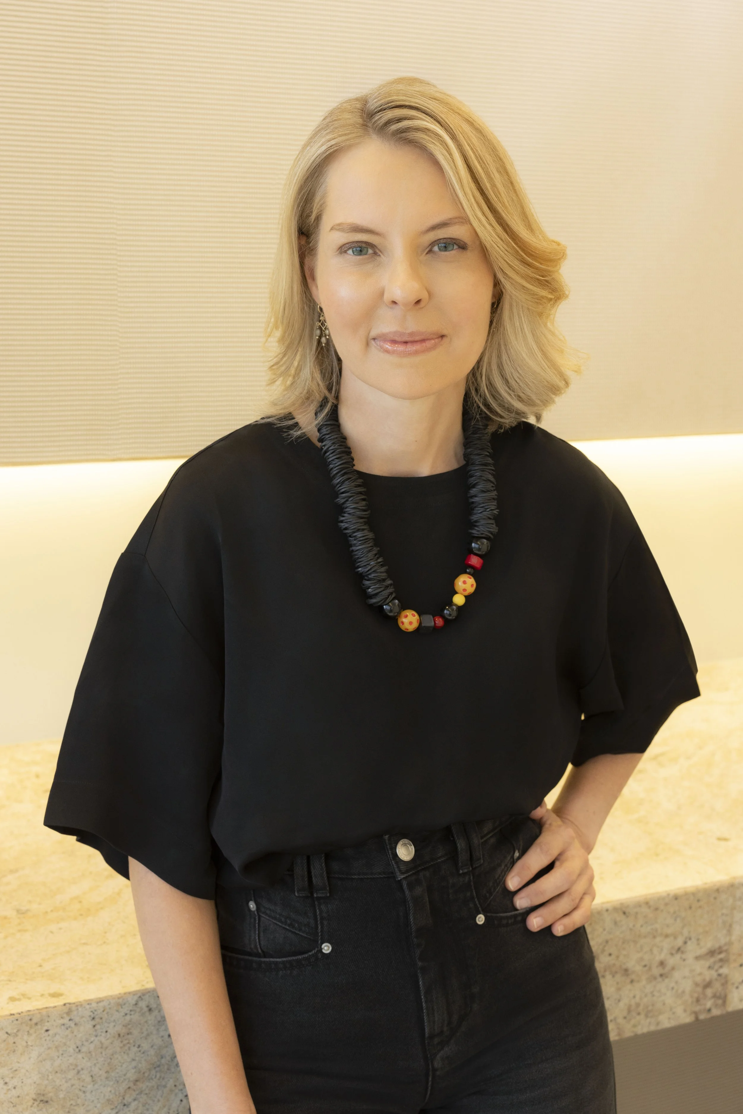 A woman with blonde hair and blue eyes wearing a black top, black jeans, and a colorful beaded necklace, standing indoors.