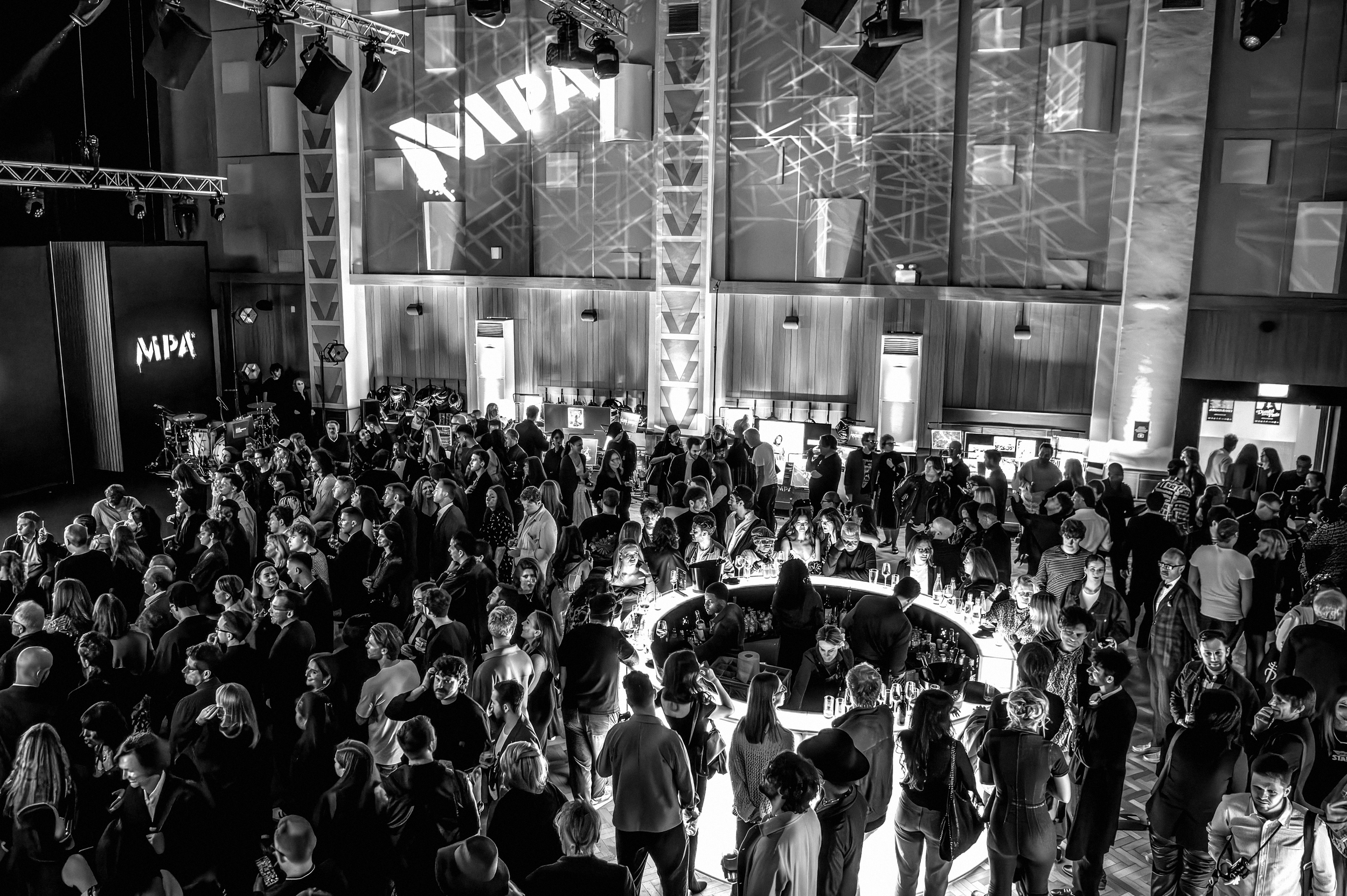 A black and white photo of a large indoor event with many people gathered, some standing and some walking, in what appears to be a conference or festival setting with a stage, lighting, and a bar area visible.