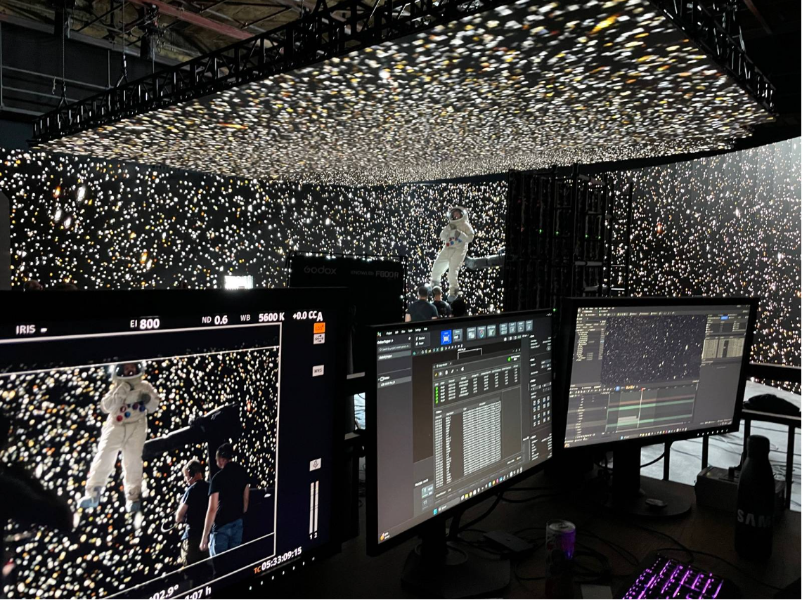 A large screen with an astronaut in a white spacesuit floating in a starry space background, with two additional monitors in the foreground showing video editing software, and controllers and equipment on a desk.