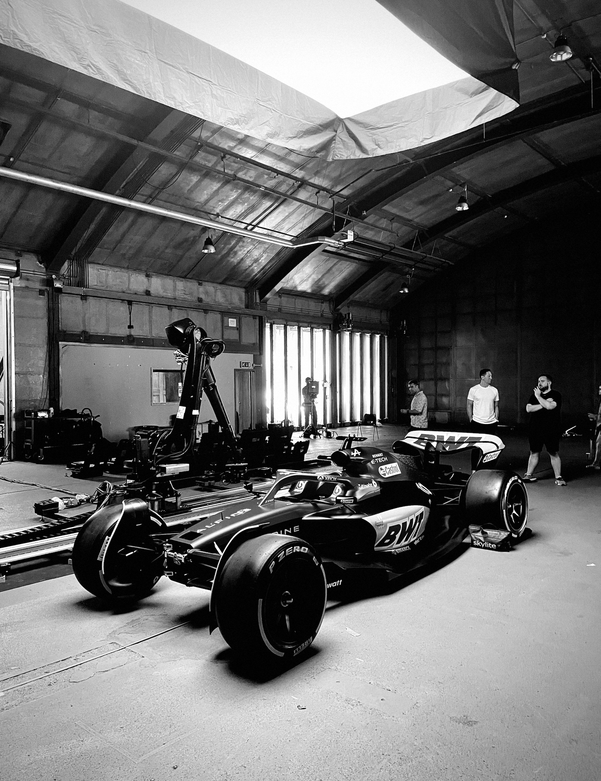 Black and white photo of an indoor garage or workshop with a Formula 1 race car on a ramp, a pole, and three men standing and conversing in the background.