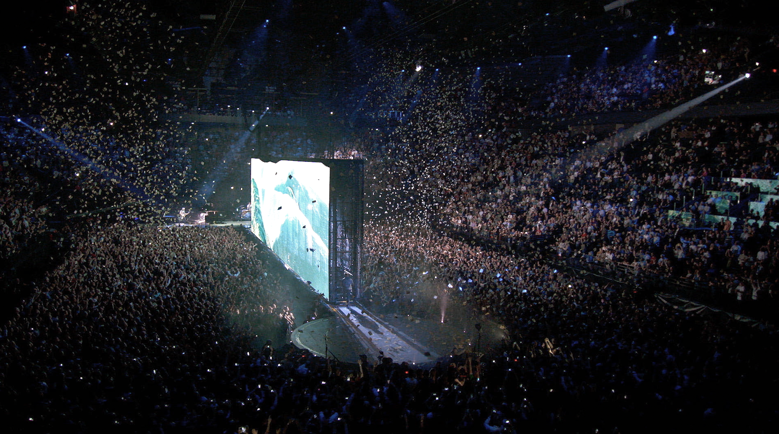 A packed indoor concert venue with a large crowd, stage, and multiple screens displaying visuals. Confetti is falling from above as blue and white lights illuminate the scene.