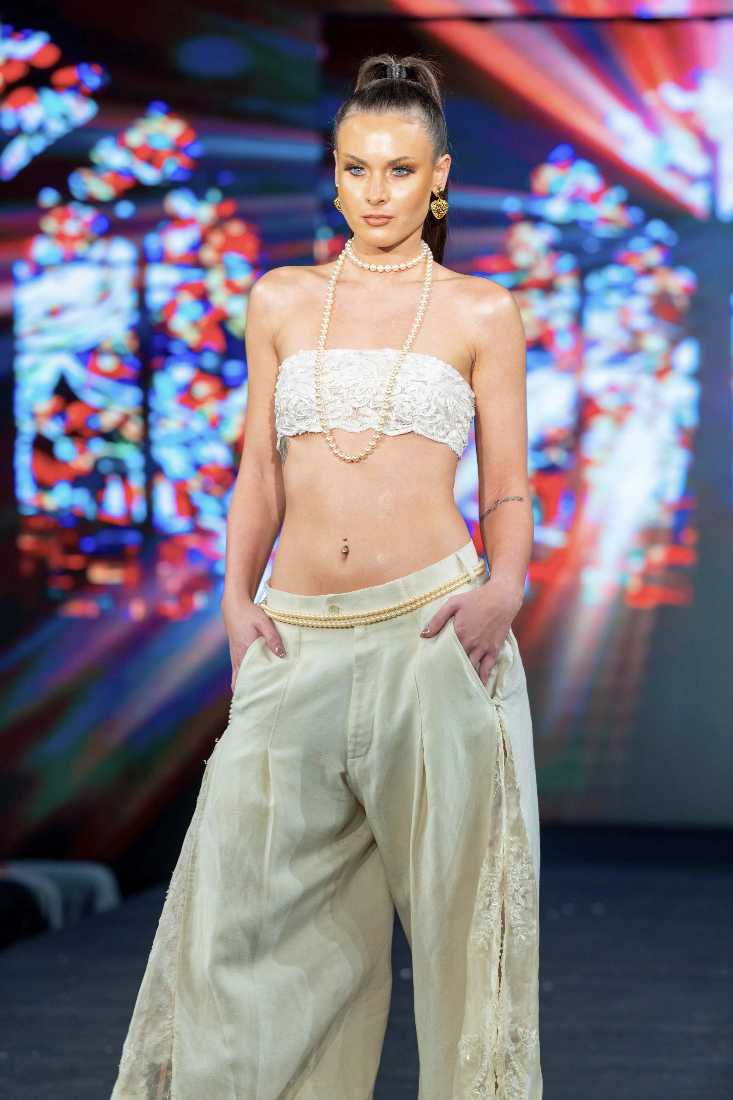 A woman with long brown hair tied in a high ponytail, wearing a white lace strapless top, cream colored wide-leg pants with embroidery details, layered pearl necklaces, and gold earrings, posing in a setting with colorful, blurred LED lights in the background.