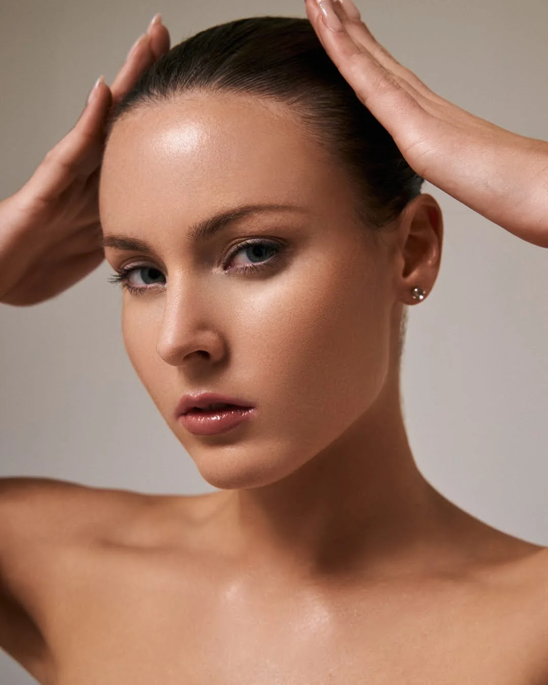 Close-up of a woman with smooth skin, striking blue eyes, and glossy lips, holding her hands on her head against a neutral background.
