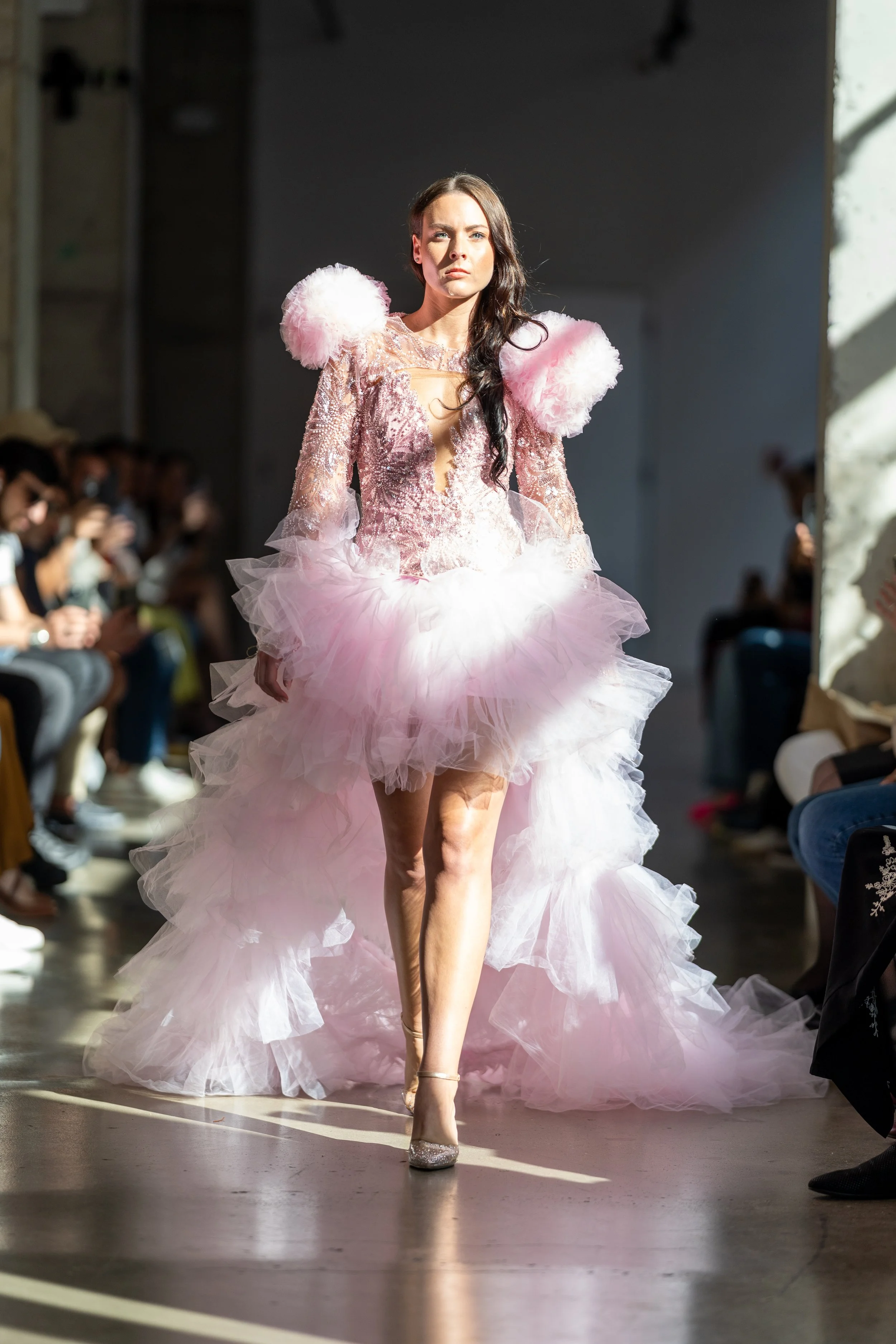 A female model walks down the runway in a pink, ruffled gown with large, fluffy shoulder details, silver high heels, and dark wavy hair, during a fashion show with seated audience on either side.