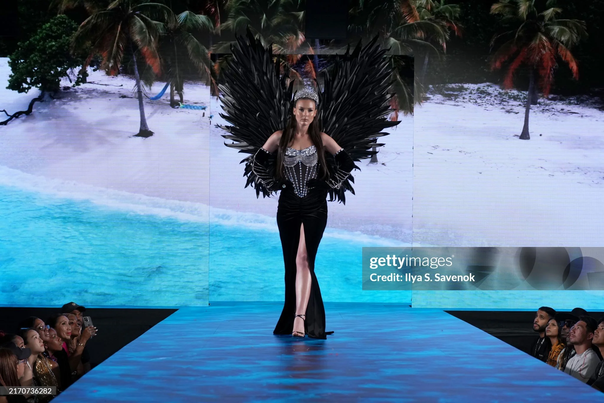 A woman walking the runway during a fashion show, wearing a black gown and elaborate black feathered and jeweled headdress, against a backdrop of a beach scene with trees and water.