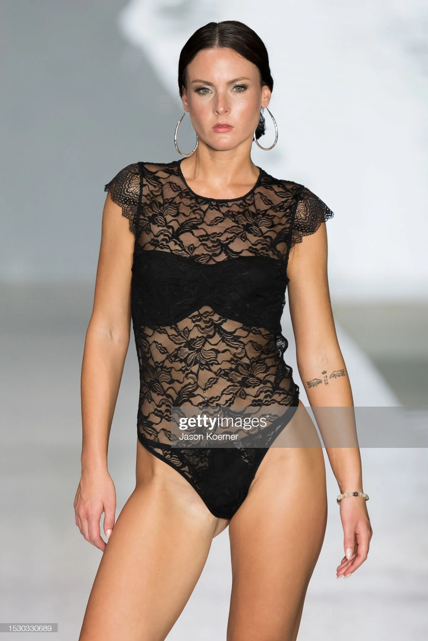 A woman with dark hair styled in a low bun runway modeling in black lace lingerie, silver hoop earrings, and a beaded bracelet on her right wrist, against a plain light gray background.