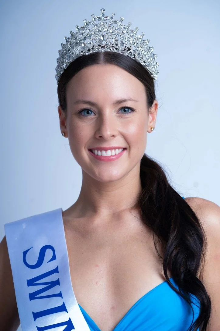 A woman wearing a crown and sash, smiling, with dark hair tied to one side, against a light background.
