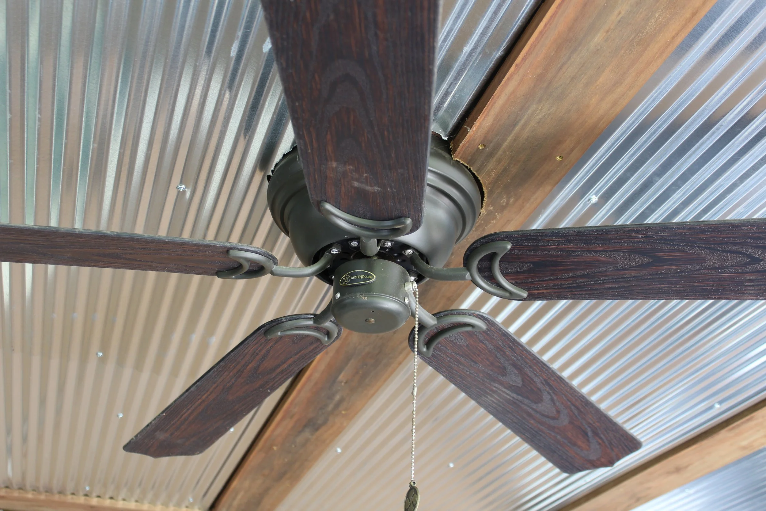 Close-up of a ceiling fan with four wooden blades and a central motor, mounted on a ceiling with a metallic, ribbed surface.
