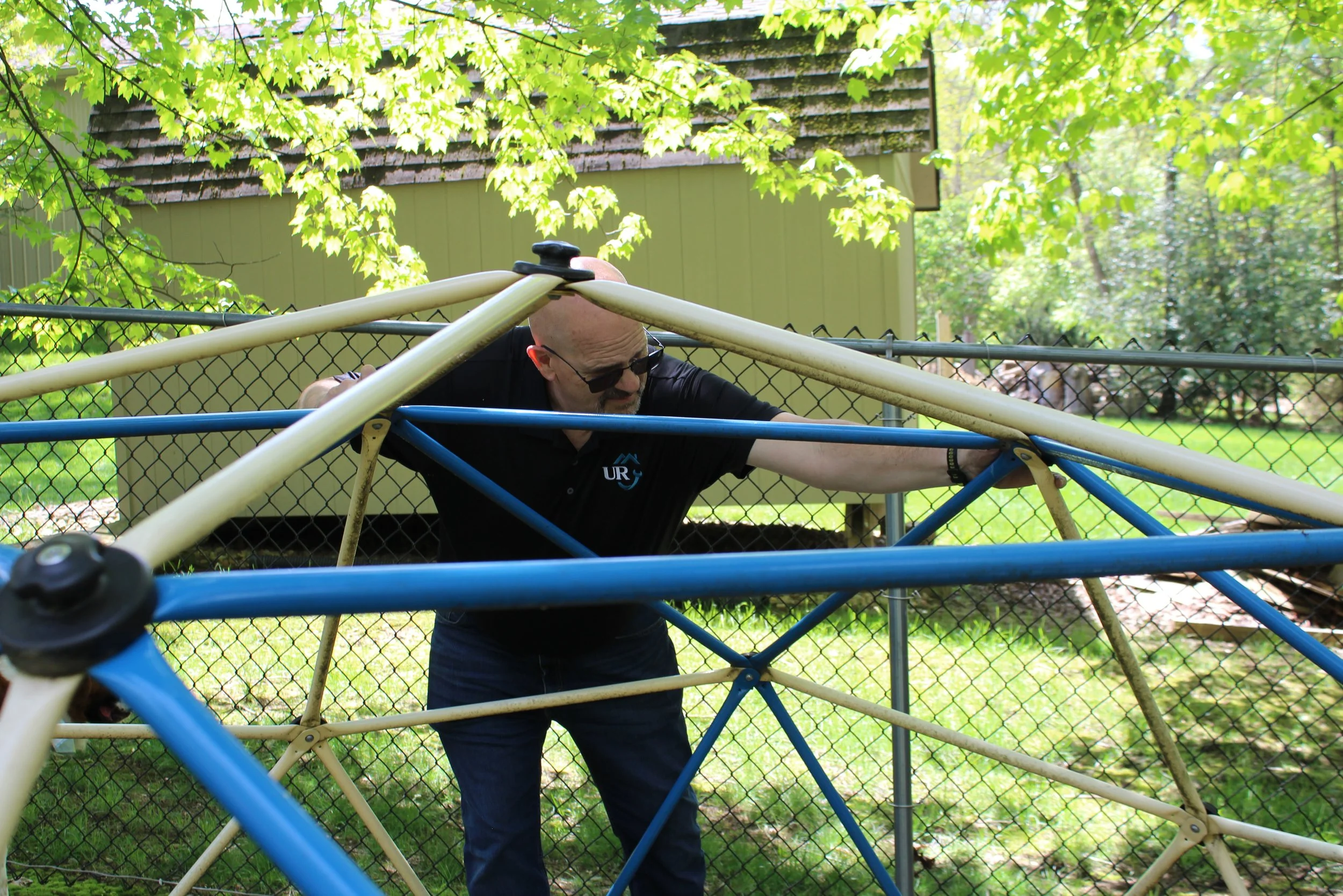 A man is fixing a playground structure made of blue, white, and beige metal bars outdoors.