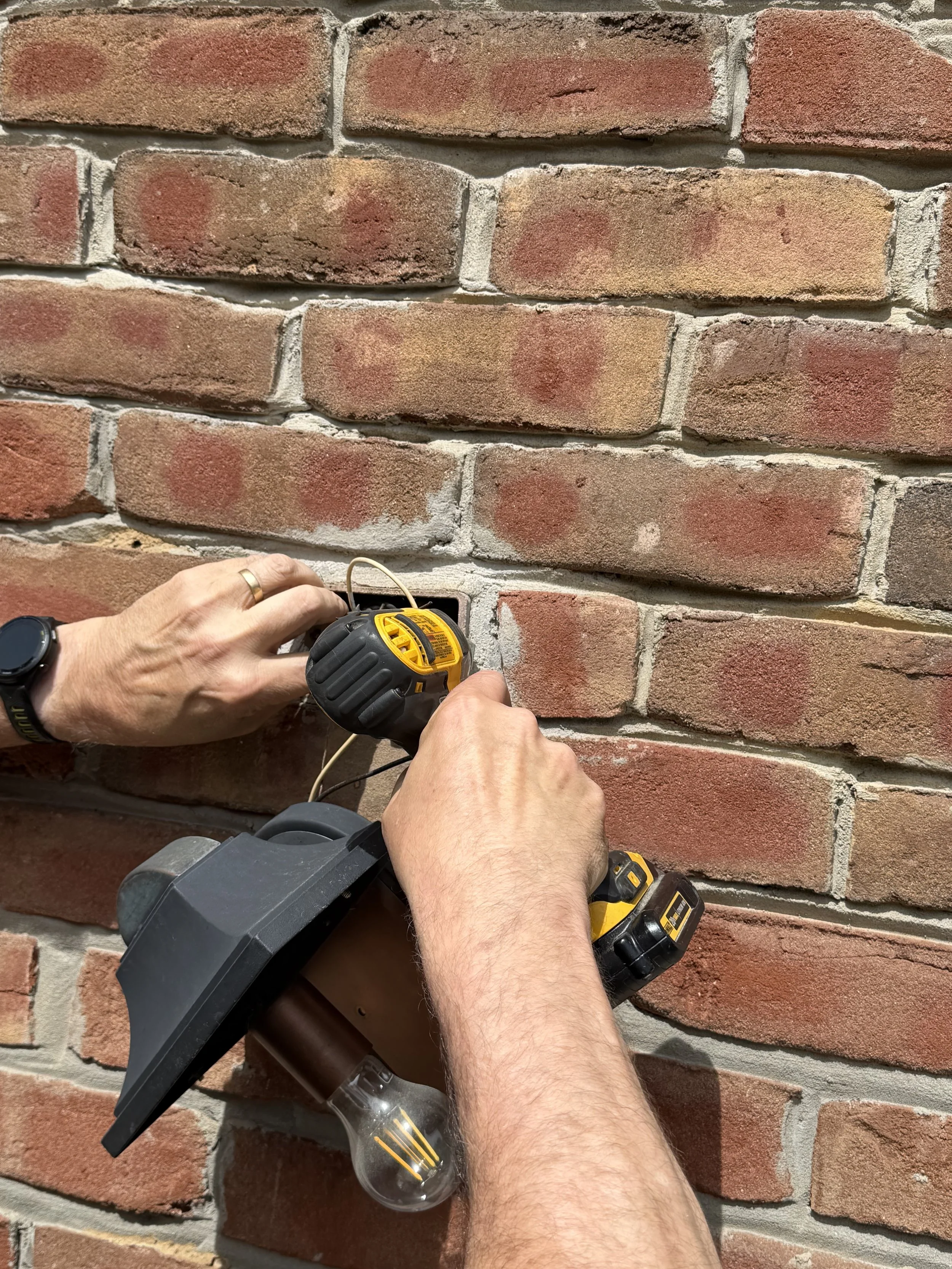 A person's hands holding a cordless drill near a brick wall, with the drill pointed downward.