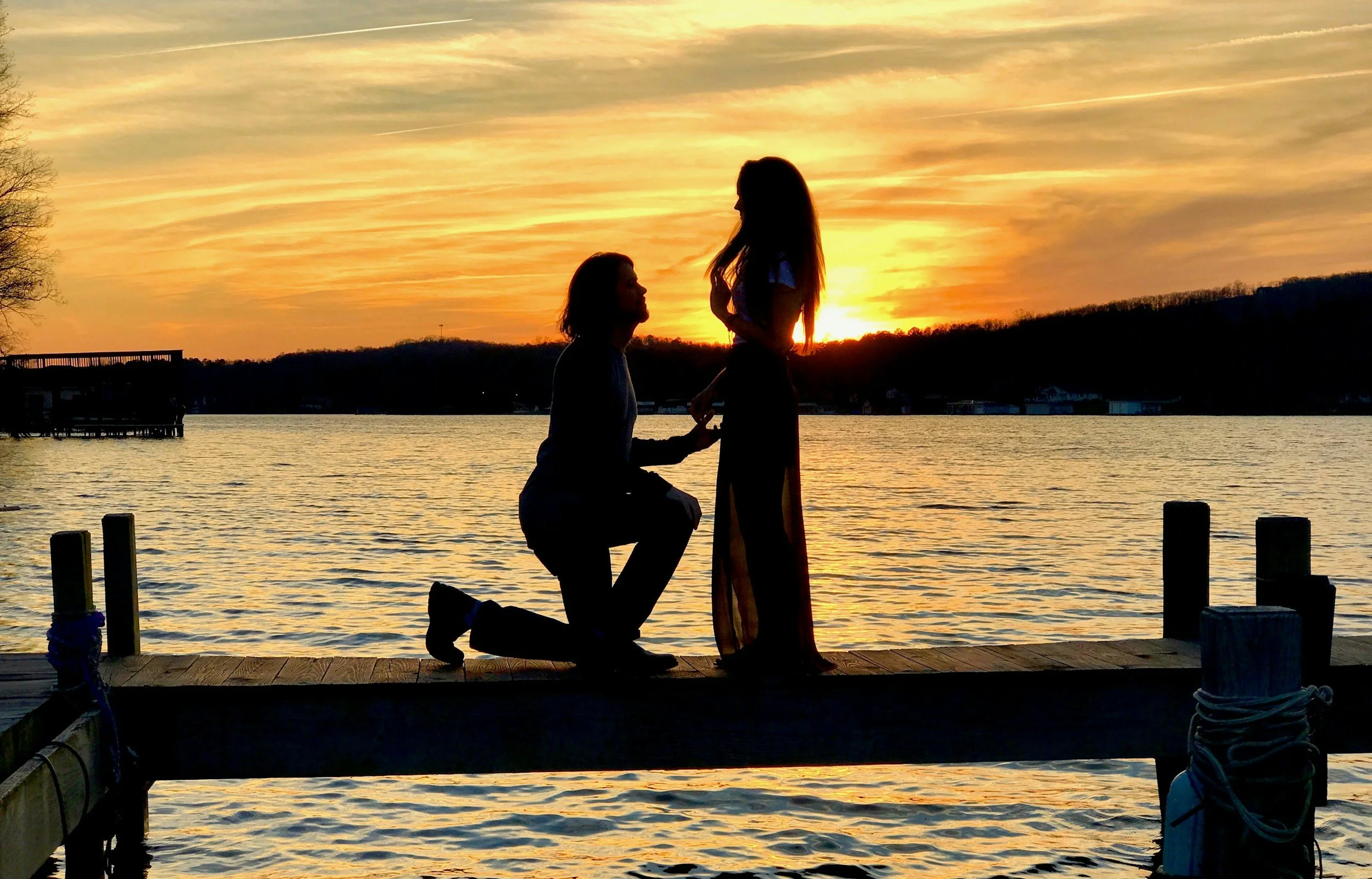 A couple at sunset by a lake, with the man kneeling on one knee holding the woman's hand, proposing. The scene is silhouetted against the colorful sky.