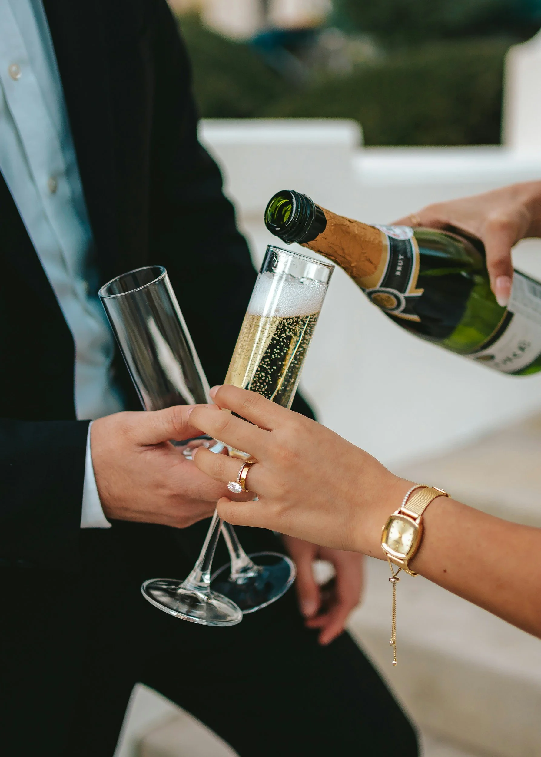 A person in a dark suit is pouring champagne from a bottle into a champagne flute held by a woman with jewelry, during a celebration or formal occasion.