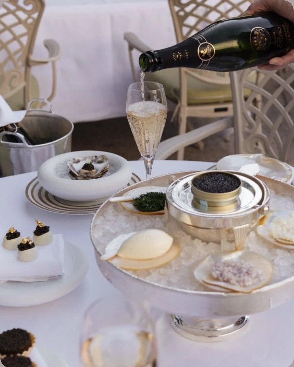 A table set with various luxury foods and a person pouring champagne into a glass. The table includes a tray of sliced cheese, soft shellfish, small appetizers topped with caviar and gold flakes, and a bowl of black caviar on ice, all on white tablecloths with chairs in the background.