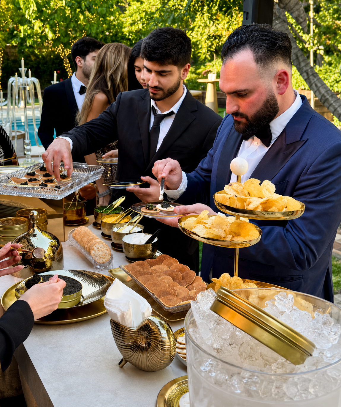 Men in tuxedos serving themselves gourmet snacks at an outdoor party with lush green trees and string lights in the background.
