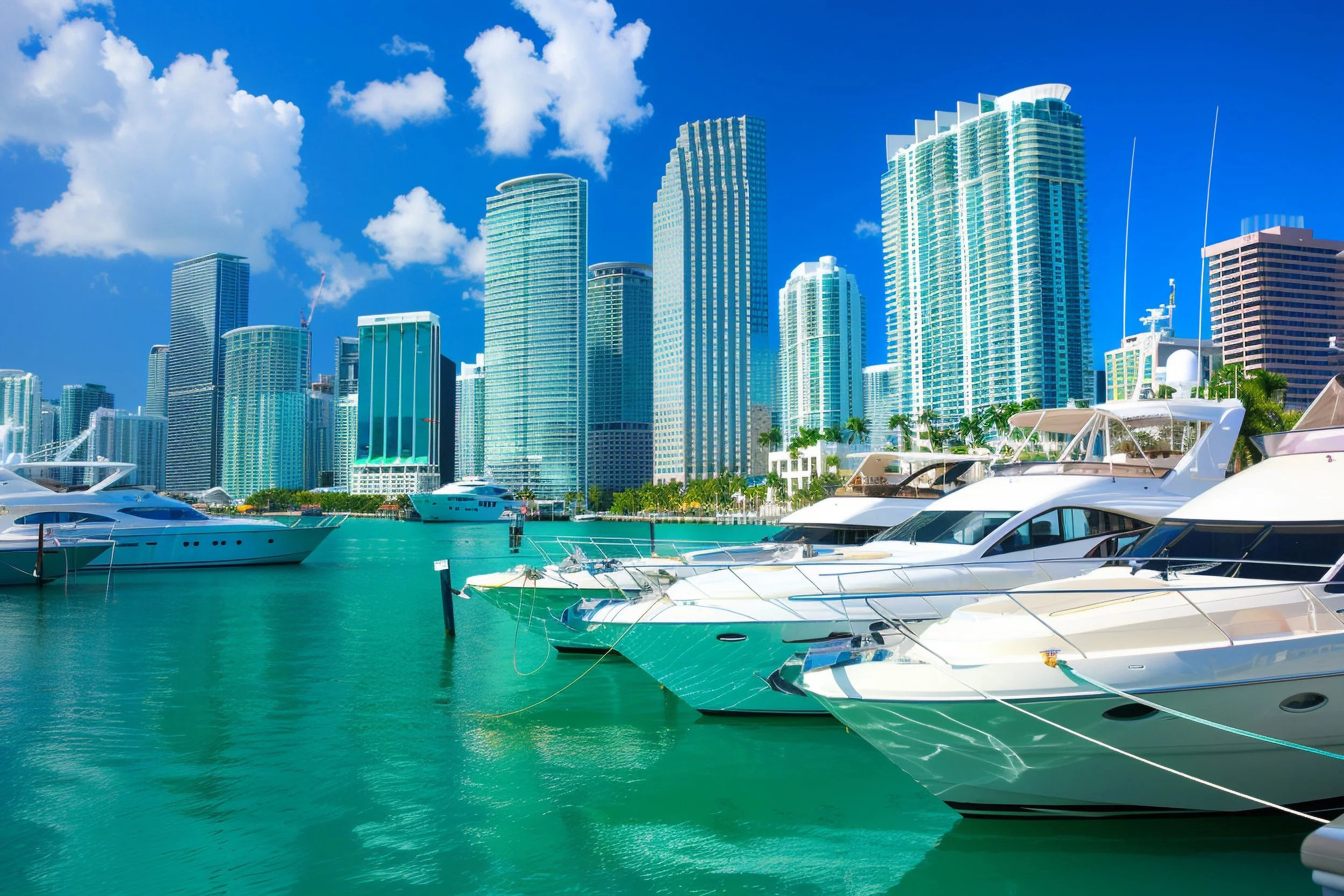 A marina with luxury yachts docked in turquoise water, with a city skyline of tall glass skyscrapers against a blue sky with white clouds in the background.