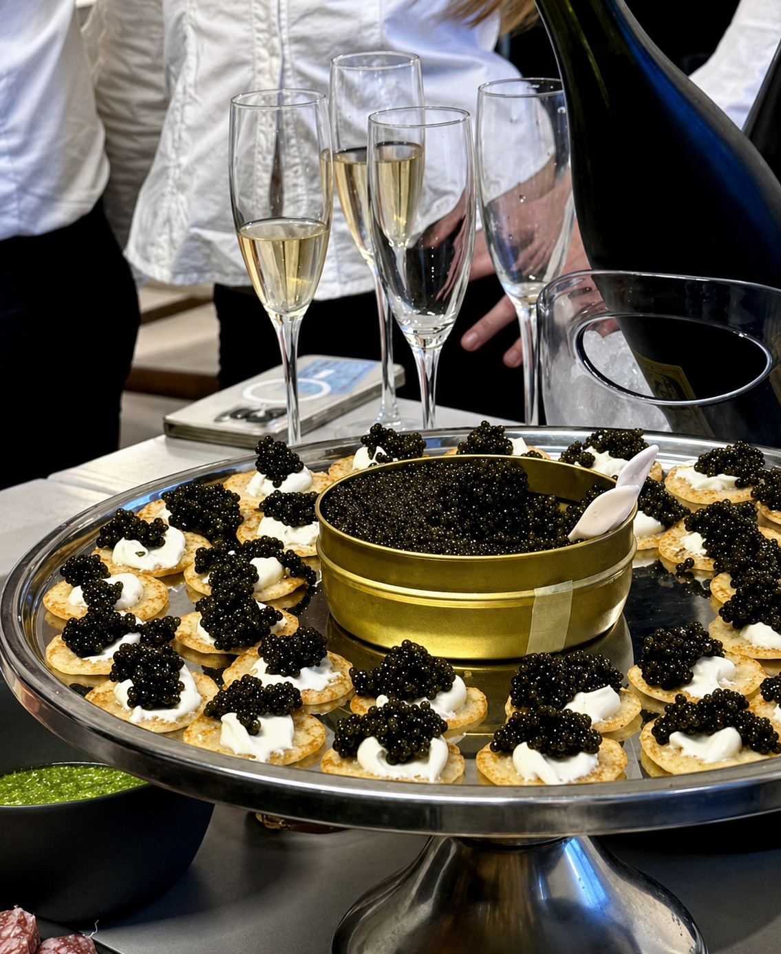 A tray of appetizers topped with black caviar and sour cream, with glasses of champagne in the background at a social gathering.
