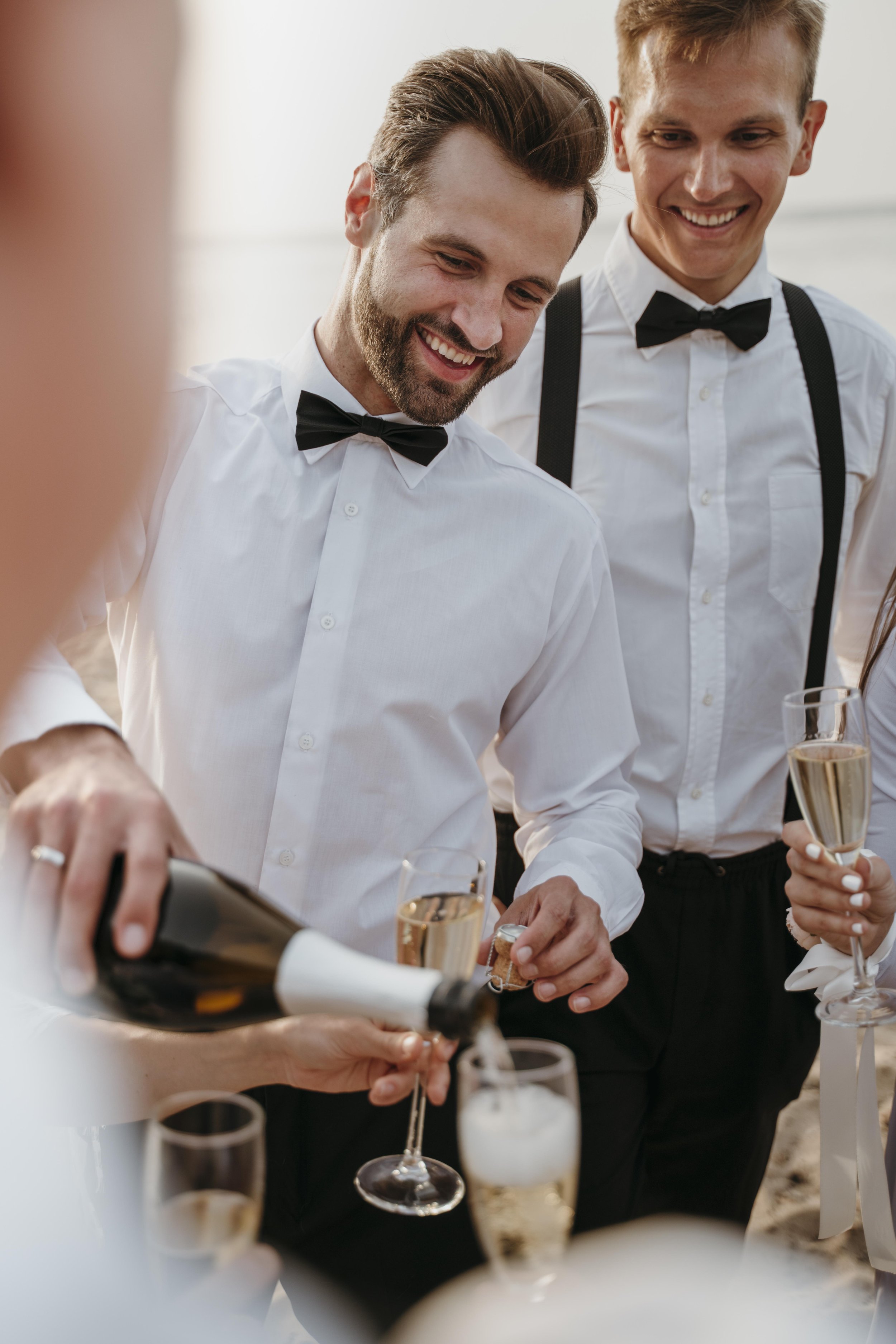 Two men wearing white shirts and black bow ties are smiling and enjoying a celebration, with glasses of champagne in their hands and a person pouring wine or champagne in the foreground.