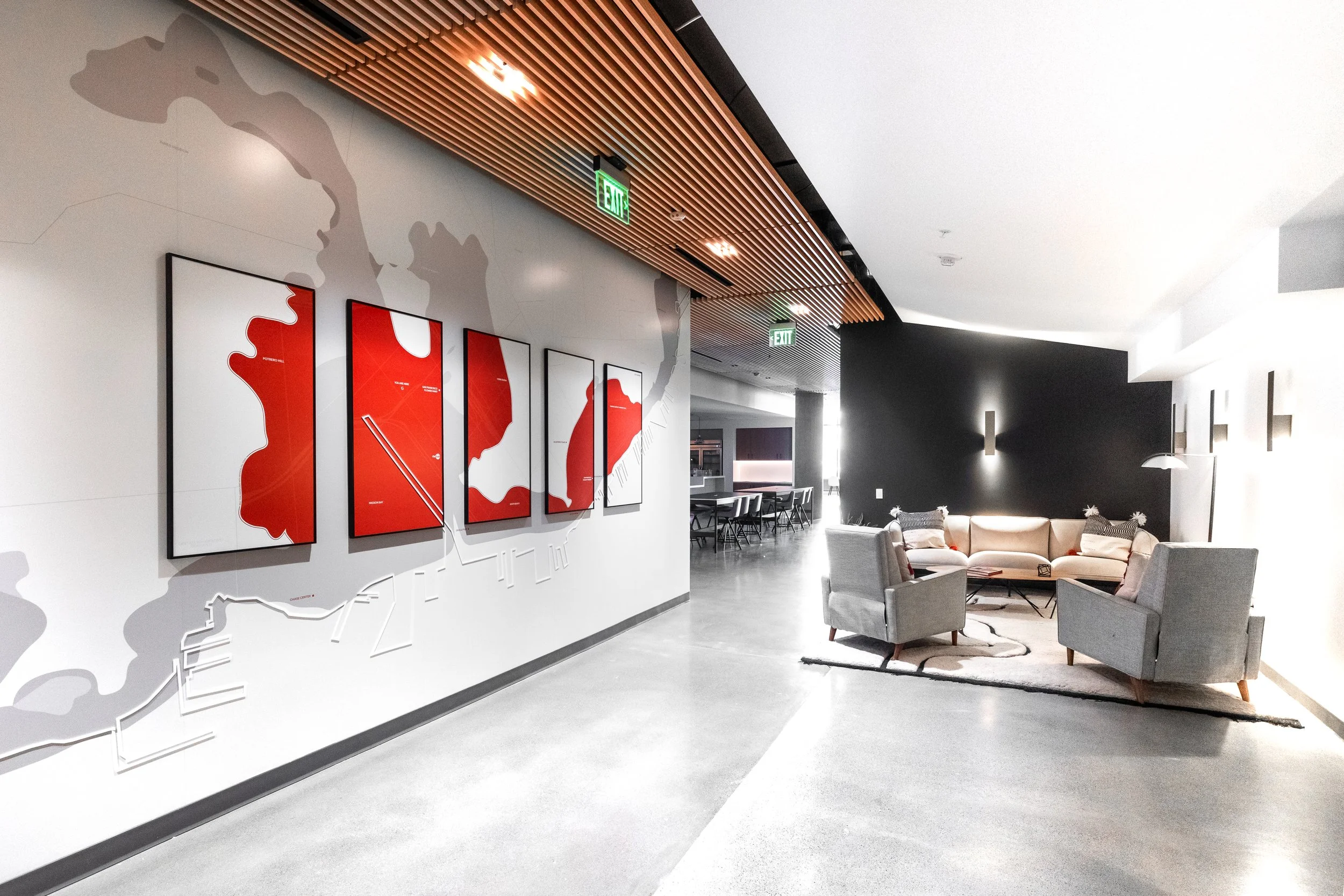 Modern lobby or reception area with a white wall featuring red map art and a partial world map, wooden slatted ceiling, gray flooring, and a seating area with beige and gray chairs and a round rug.