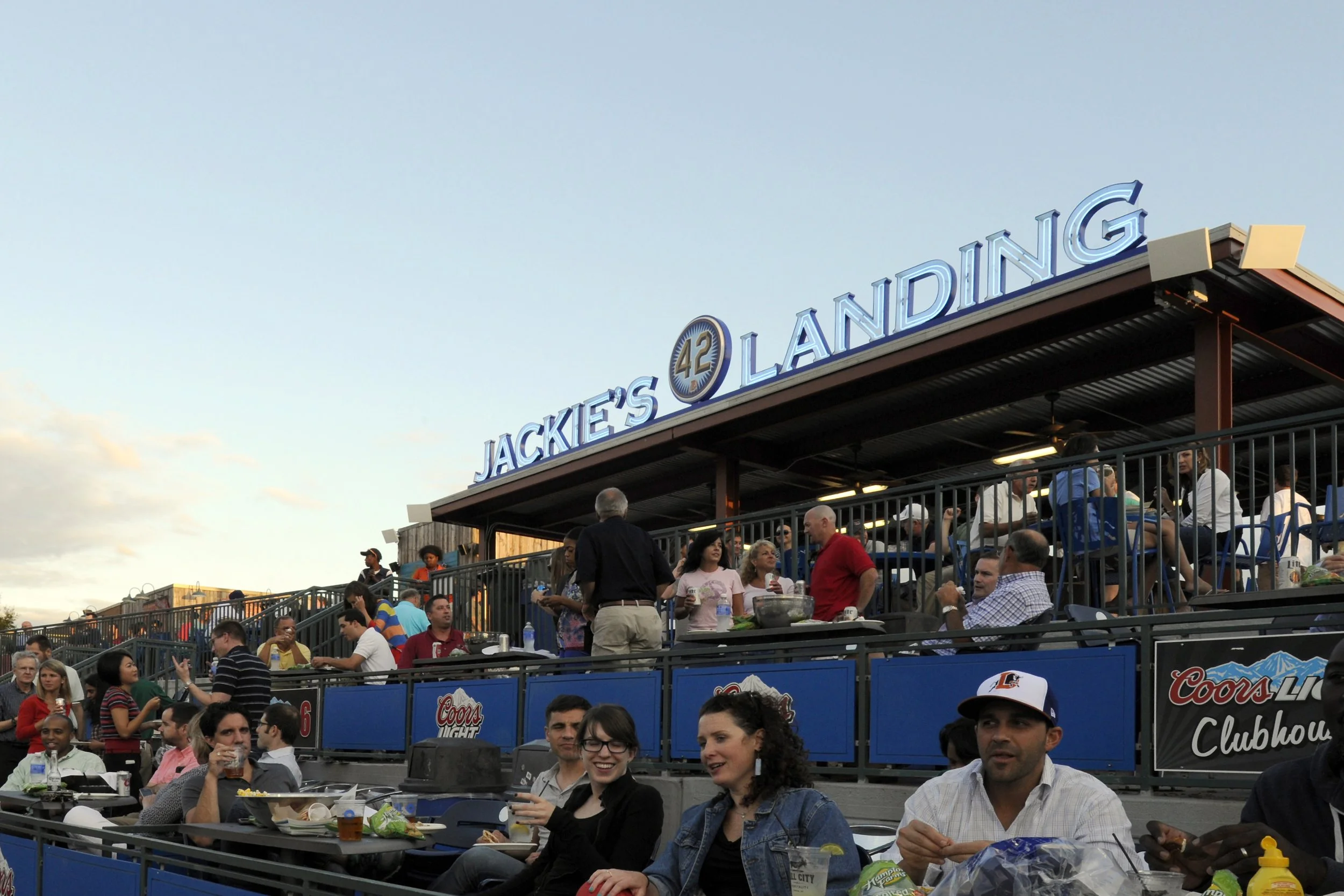 Crowd gathered at a stadium outdoor seating area during the day, with a large illuminated sign reading 'JACKIE'S LANDING' at the top, and people enjoying food and drinks.