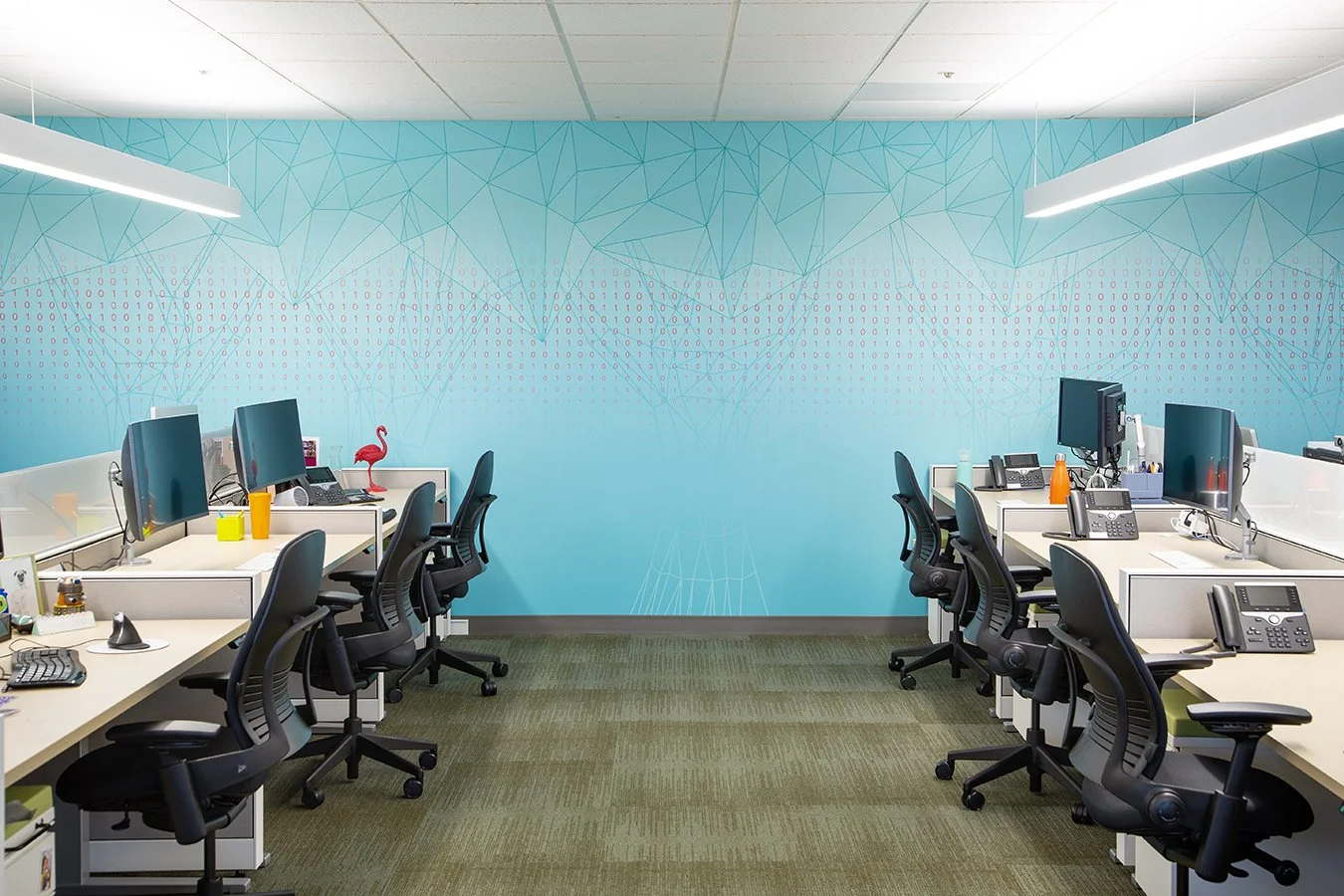 Office space with eight workstations, each with a computer monitor, phone, and office chair, arranged in two rows with a blue geometric wall design in the background.