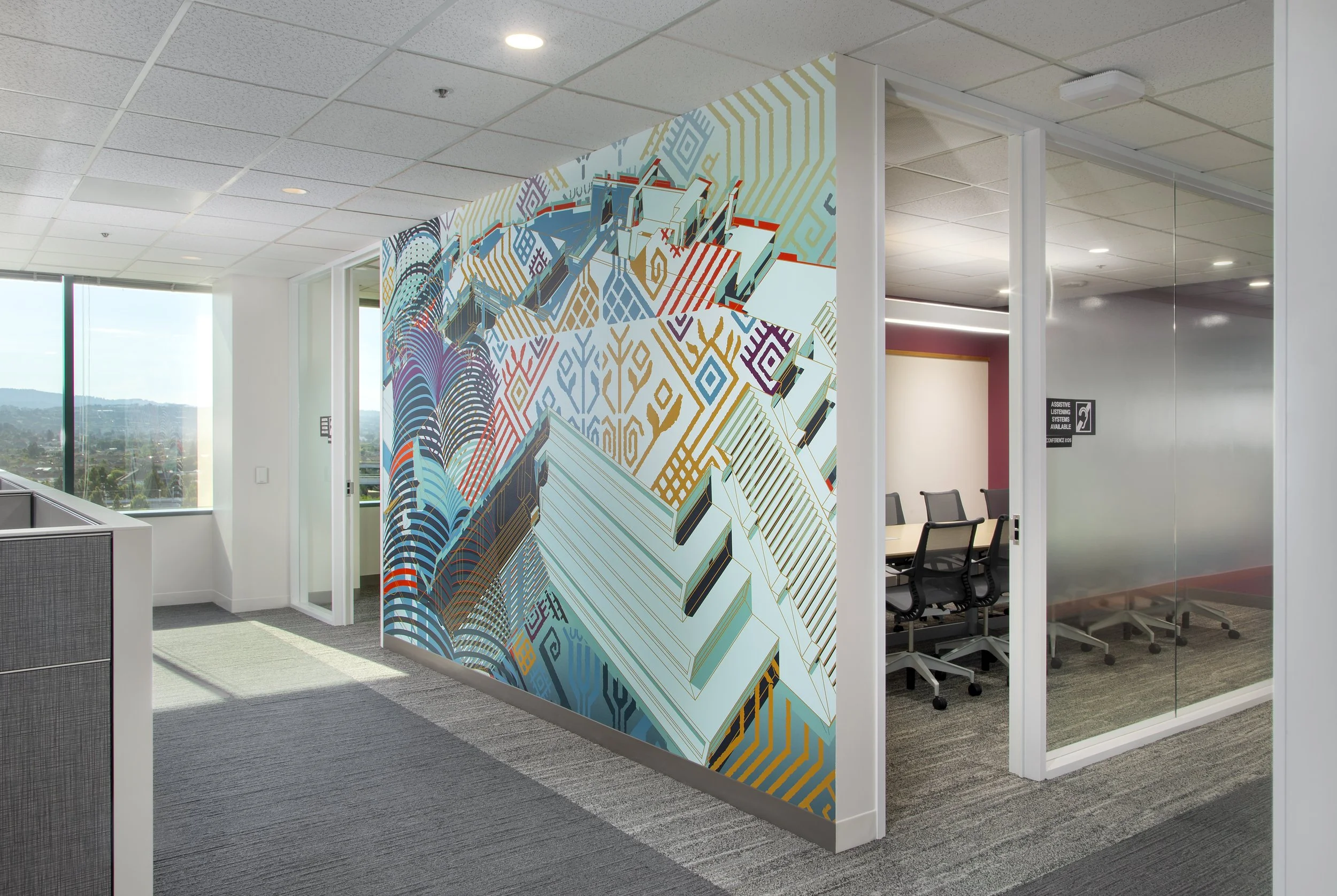 Office space with a mural wall featuring vibrant geometric and abstract patterns in blue, red, gold, and purple colors, with glass partitions and seating area visible.