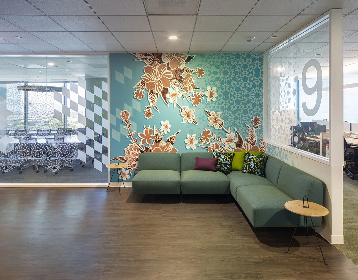 Modern office lounge area with a green sectional sofa, colorful pillows, small round wooden side tables, and a large floral mural on the wall.