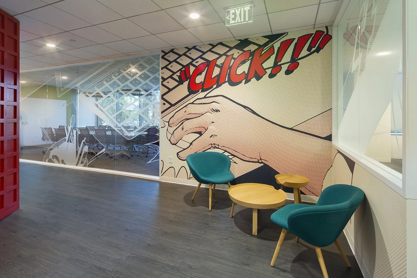 Modern office lounge area with a colorful comic book-style wall featuring a hand clicking a computer mouse and the word 'CLICK!!' in bold red letters. There are two teal armchairs, two small wooden tables, and a glass wall with office meeting rooms i
