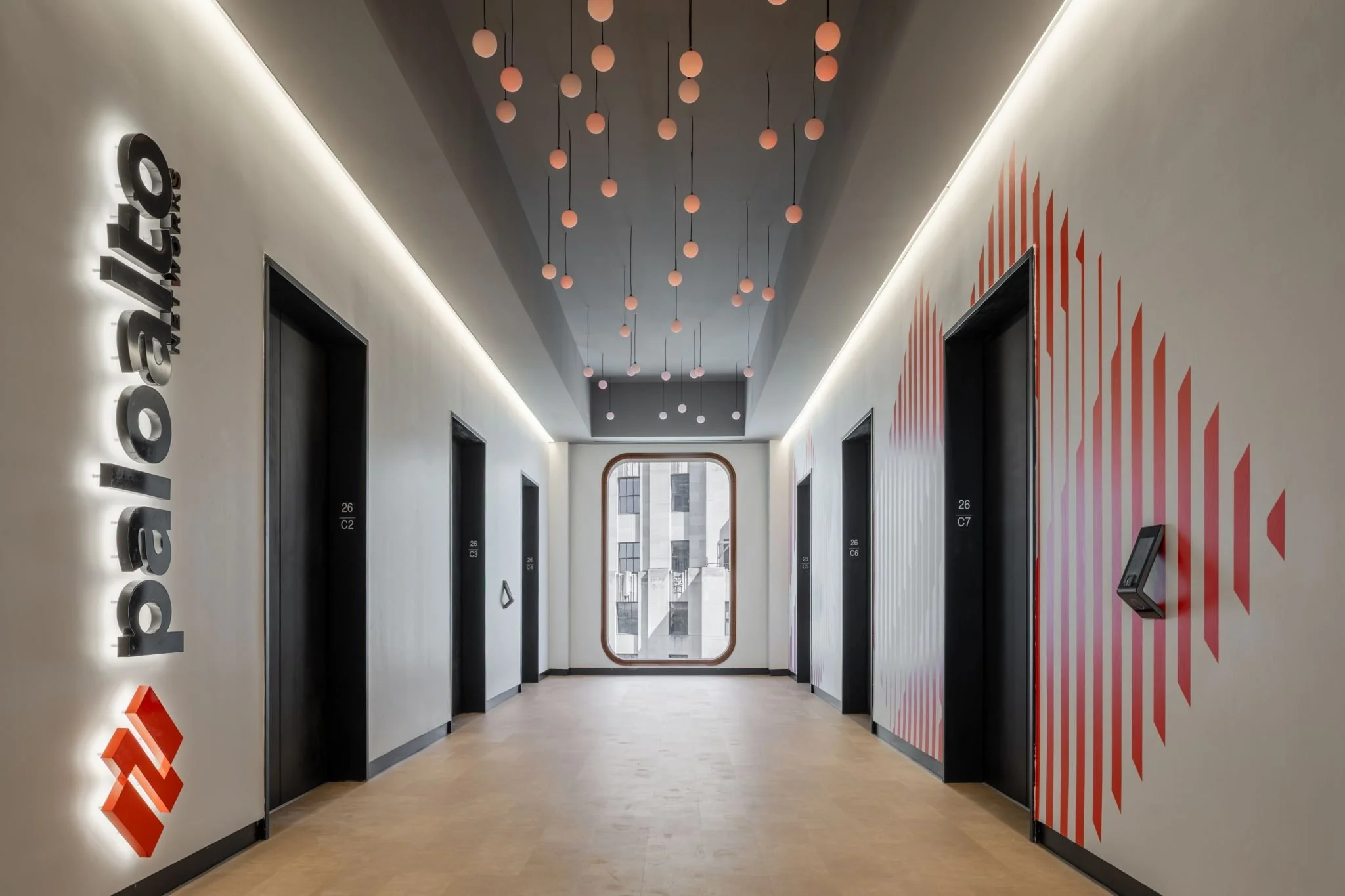 A modern elevator lobby with multiple elevator doors on either side, a decorative ceiling with hanging round light fixtures, and a large window at the end of the hallway showing city buildings outside.