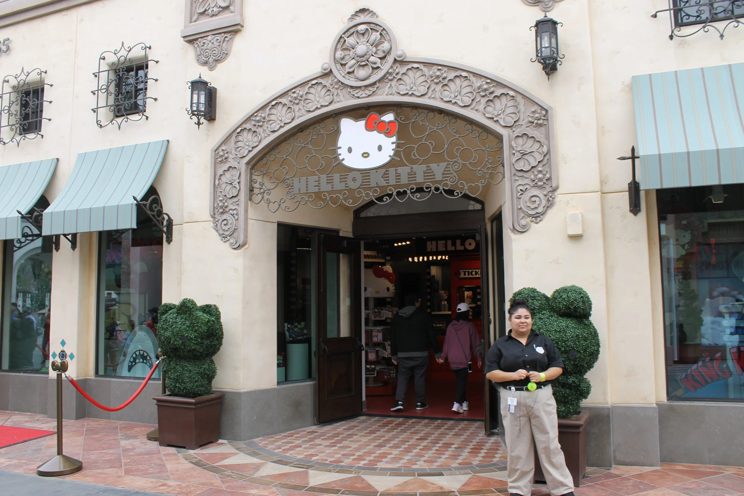 The exterior of a Hello Kitty store with an ornate archway and a Hello Kitty sign. People are entering and standing outside, with some bushes and a red rope barrier at the entrance.