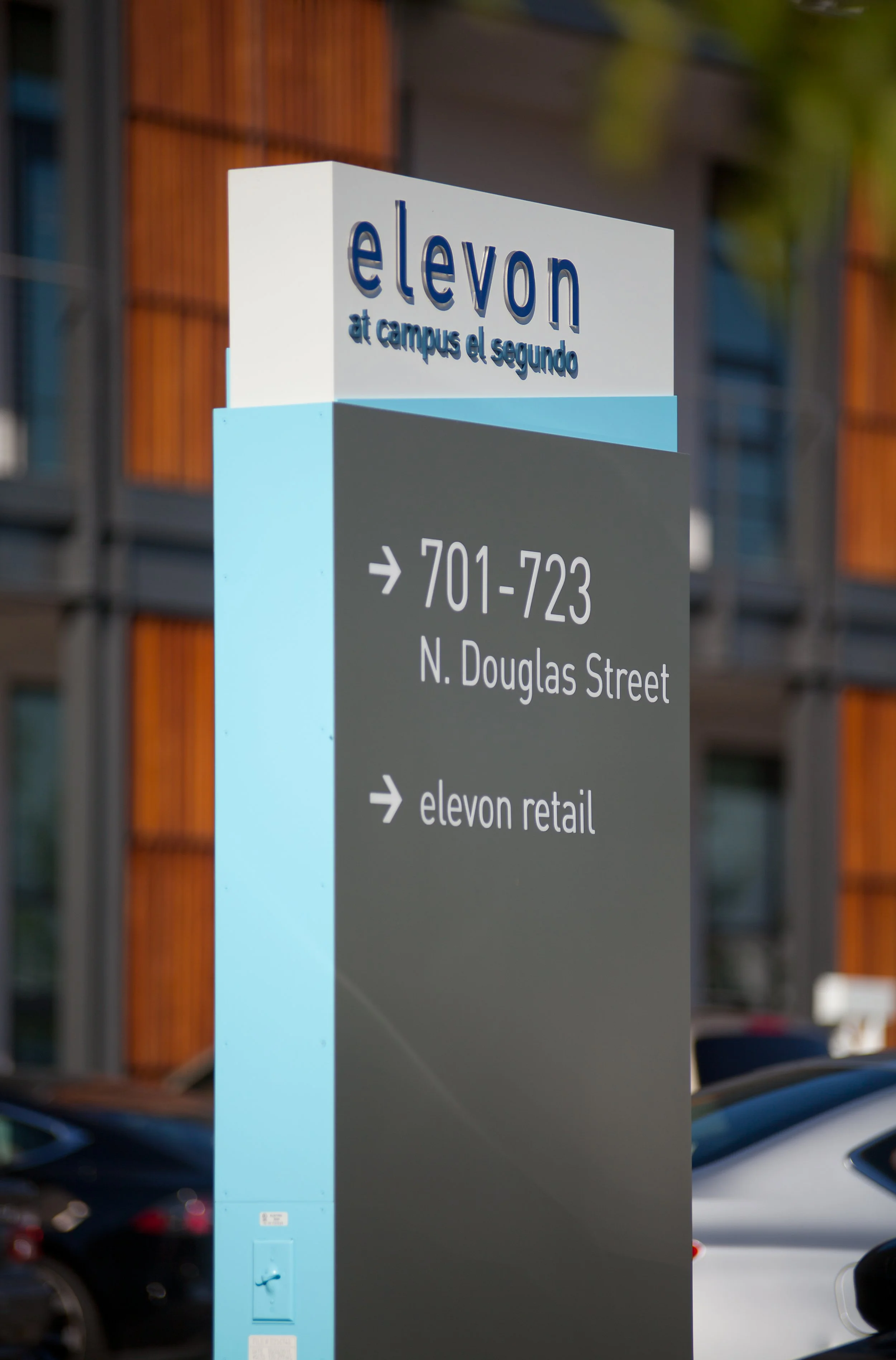 A signpost at a parking lot showing directions to Elevn at campus el Segundo, with specific parking space ranges and street name, against a background of parked cars and a building with wooden panels.