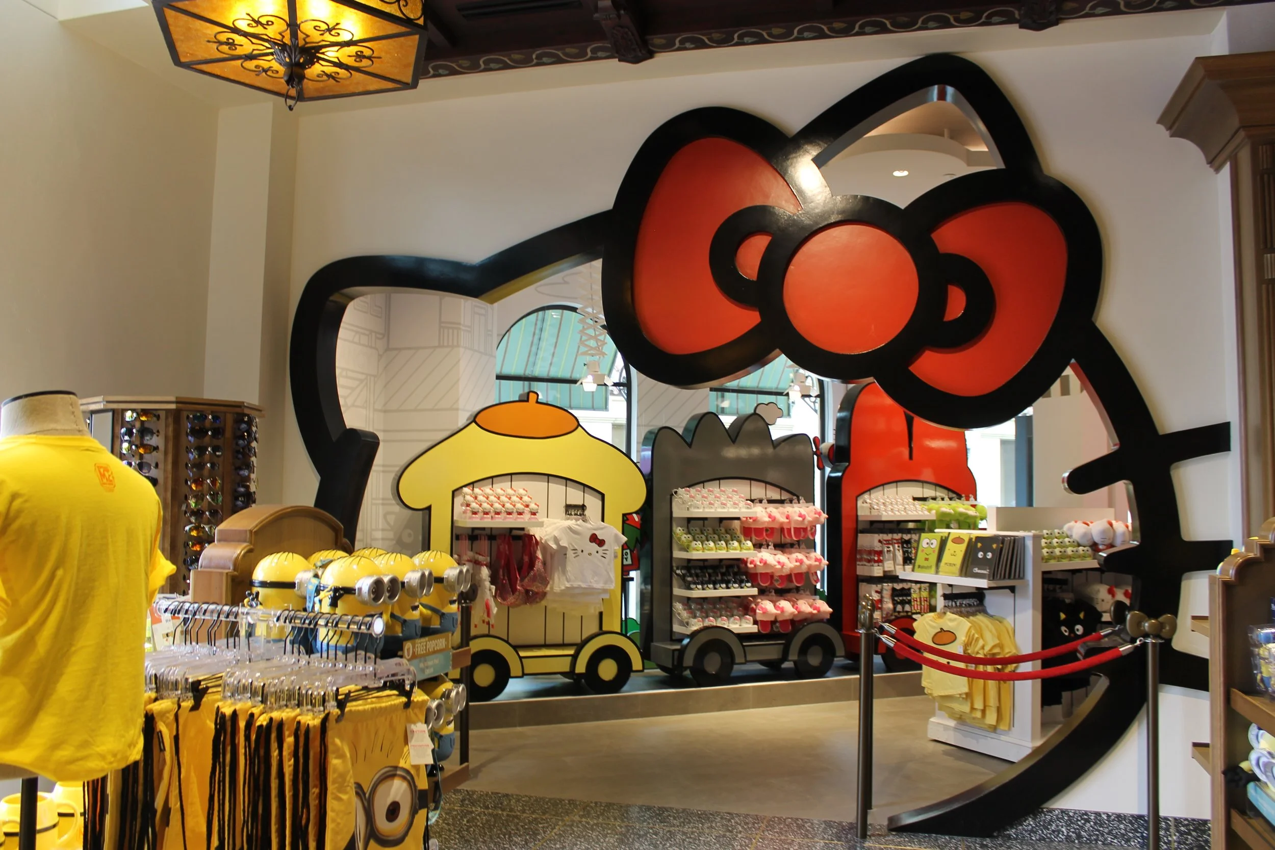 A store display with Minions-themed merchandise, including yellow shirts, plush toys, and accessories, with a large Minions graphic in the background and decorative lighting.