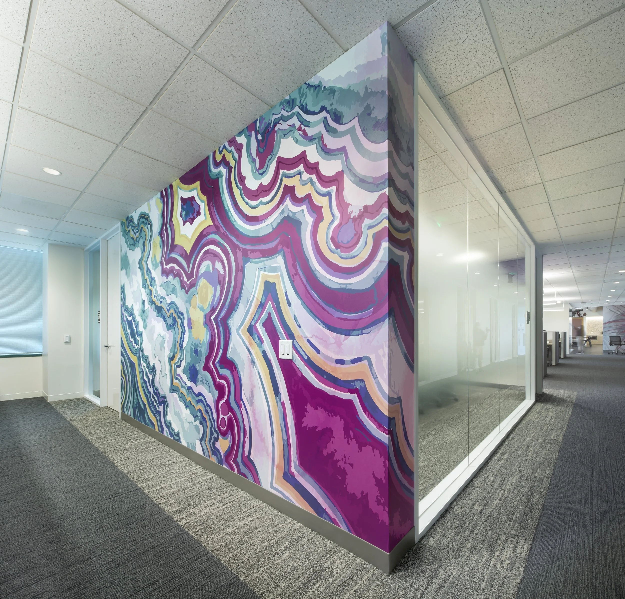 A vibrant abstract mural with swirling patterns in shades of purple, pink, blue, and white covers a corner wall in an office setting.